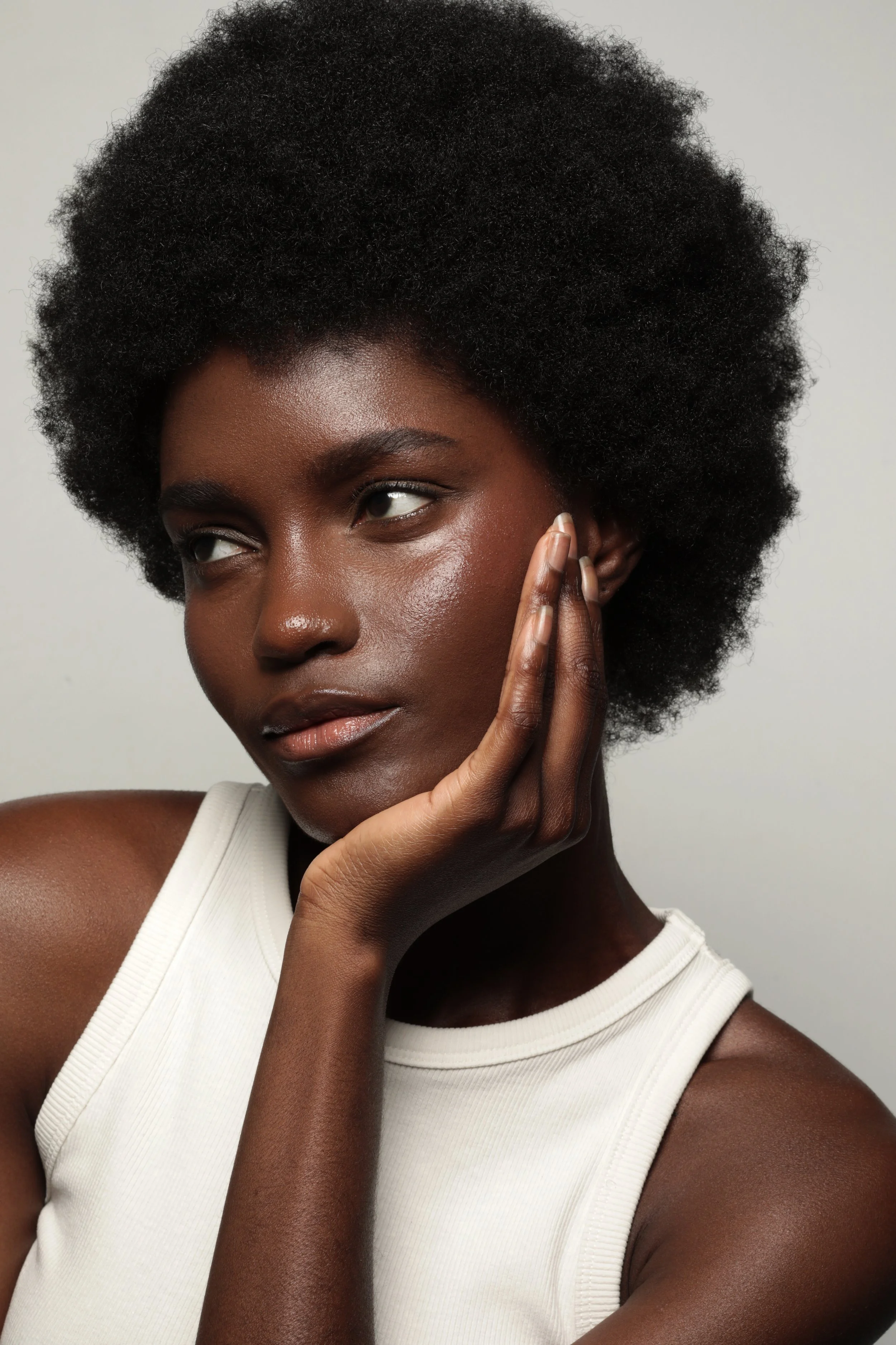 A close-up portrait of a Black woman with natural afro hair, resting her face on her hand and looking to the side, against a plain light background.