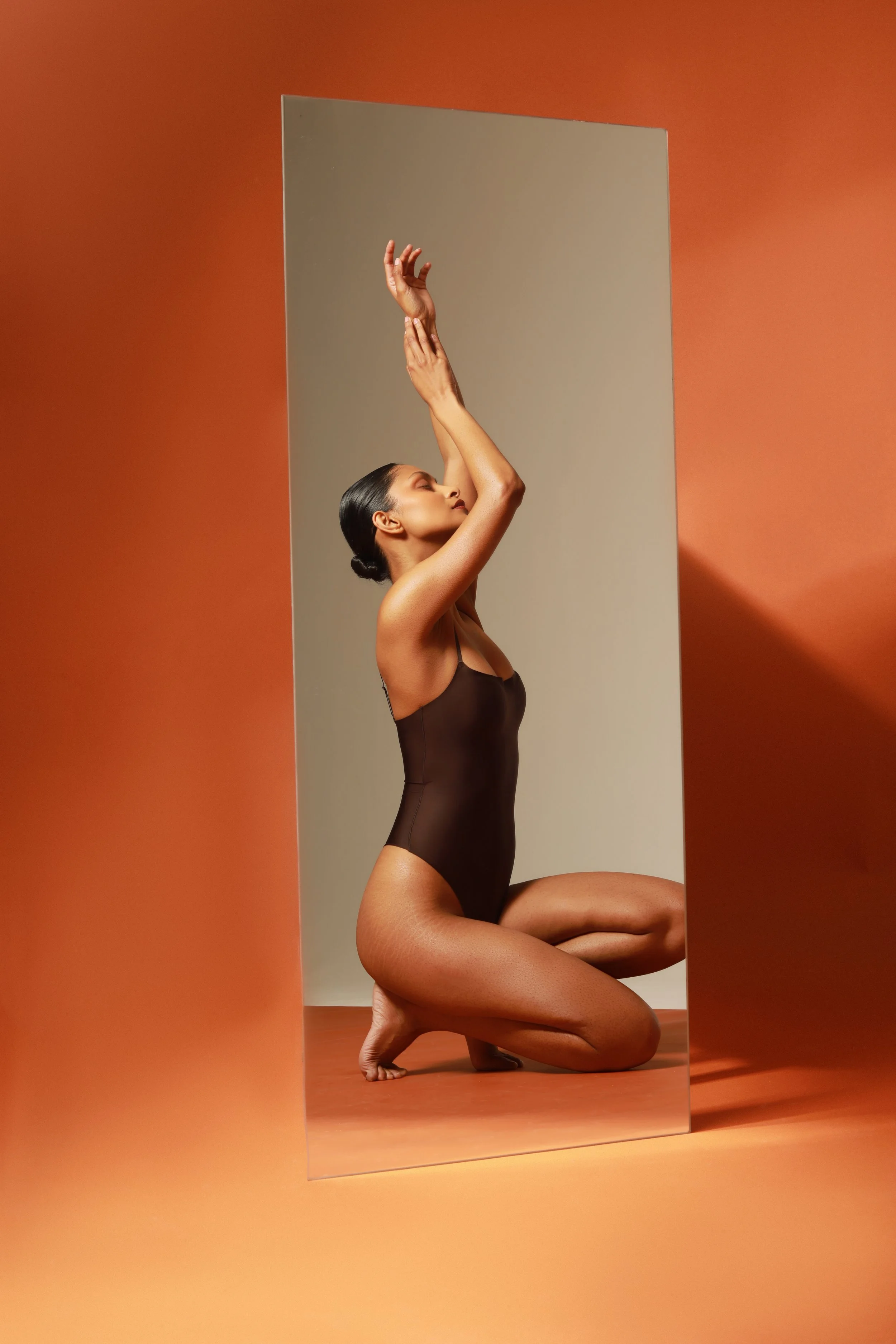 A woman with dark hair tied back, dressed in a dark bodysuit, poses on her knees with one knee bent and the other foot flat, reaching upward with her right arm, in front of a large mirror, on an orange background.