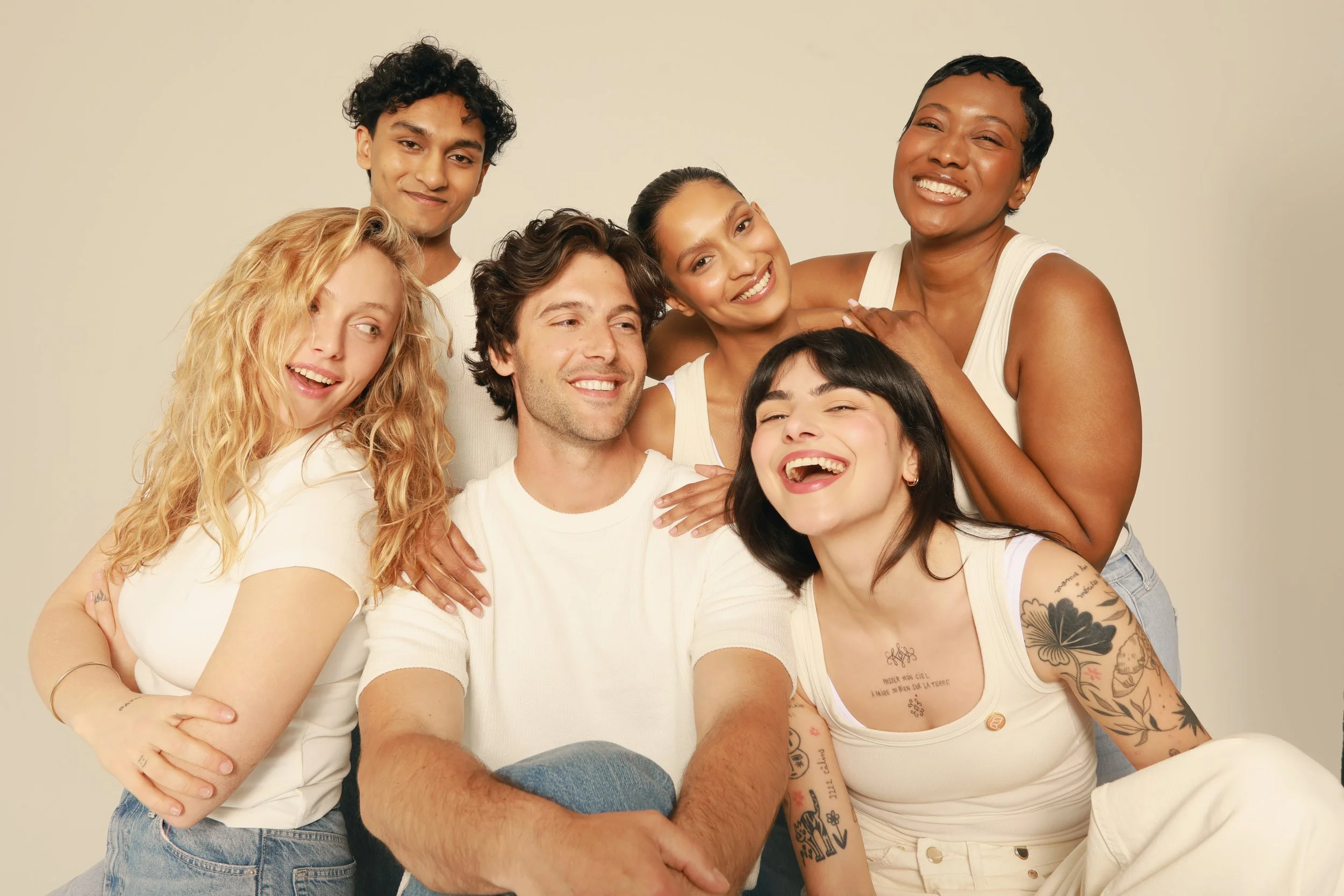 A group of six diverse people smiling and hugging against a plain beige background.