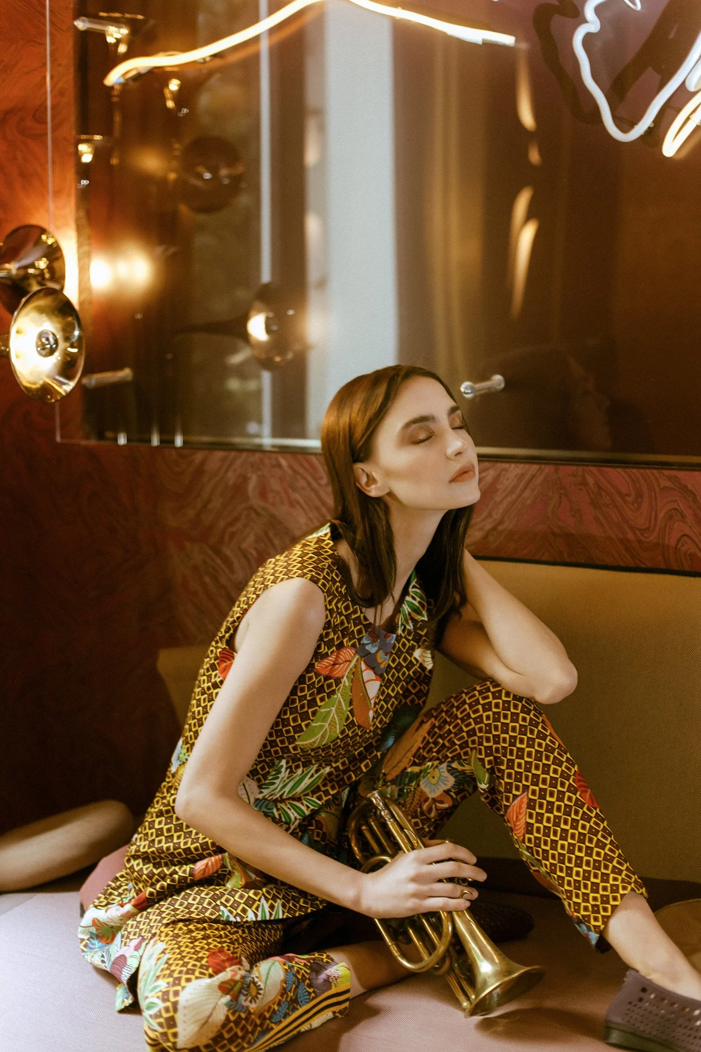 A woman with long brown hair, closed eyes, and floral-patterned clothing sitting on the floor, leaning against a wall, holding a brass trumpet in her lap, in a warmly lit room with reflective surfaces and decorative elements.
