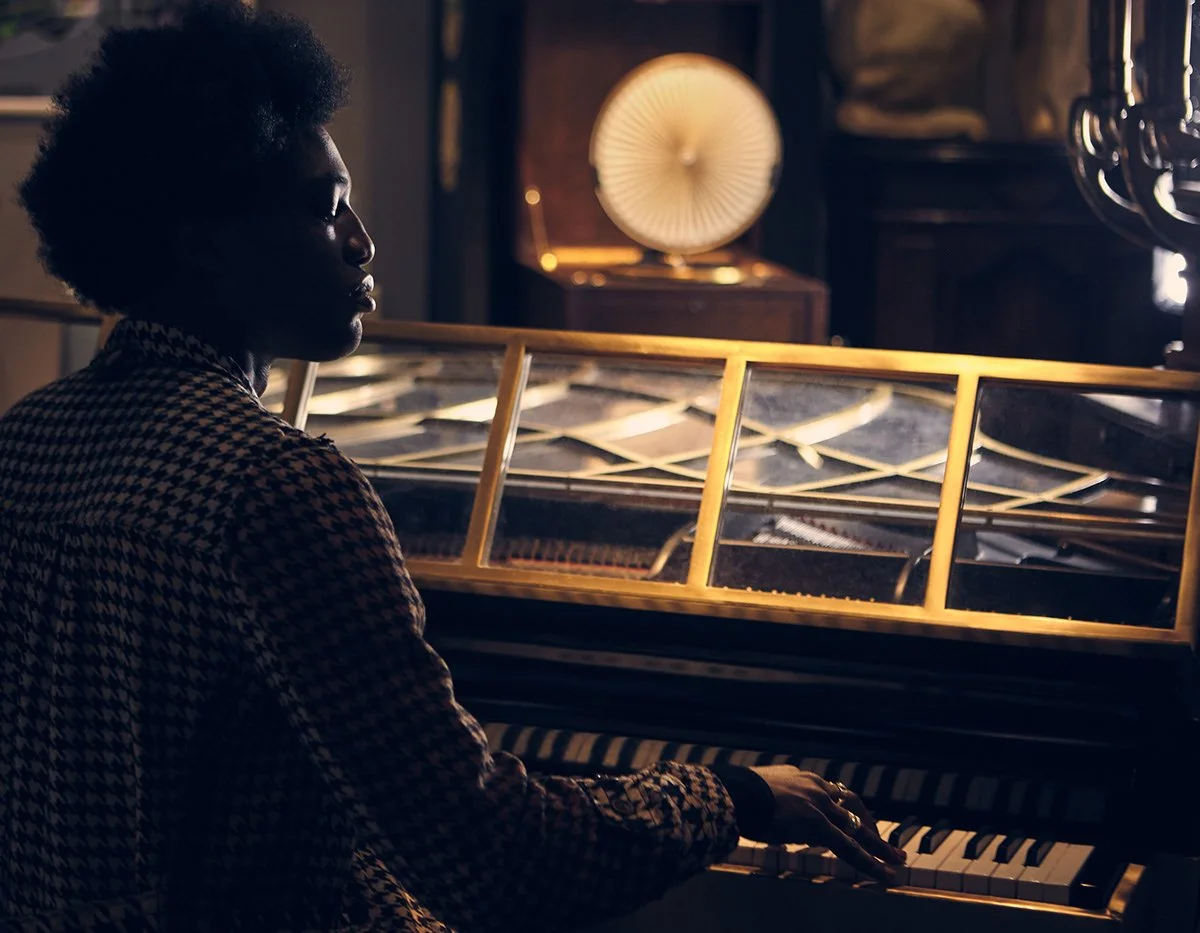 A woman playing a grand piano in a dimly lit room with warm, ambient lighting and decorative objects in the background.
