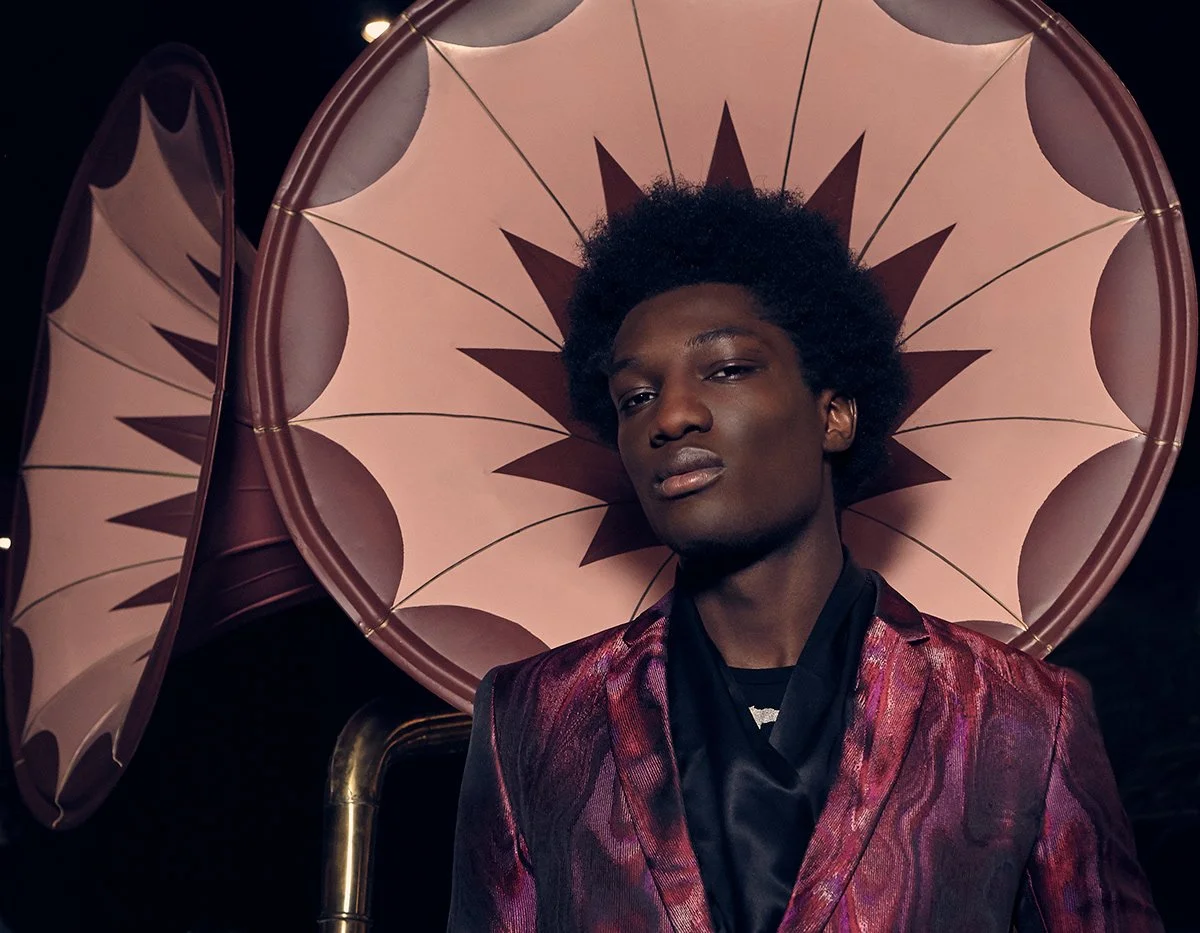 A young man with dark skin and an afro hairstyle standing in front of a large decorative pink umbrella with a star pattern.
