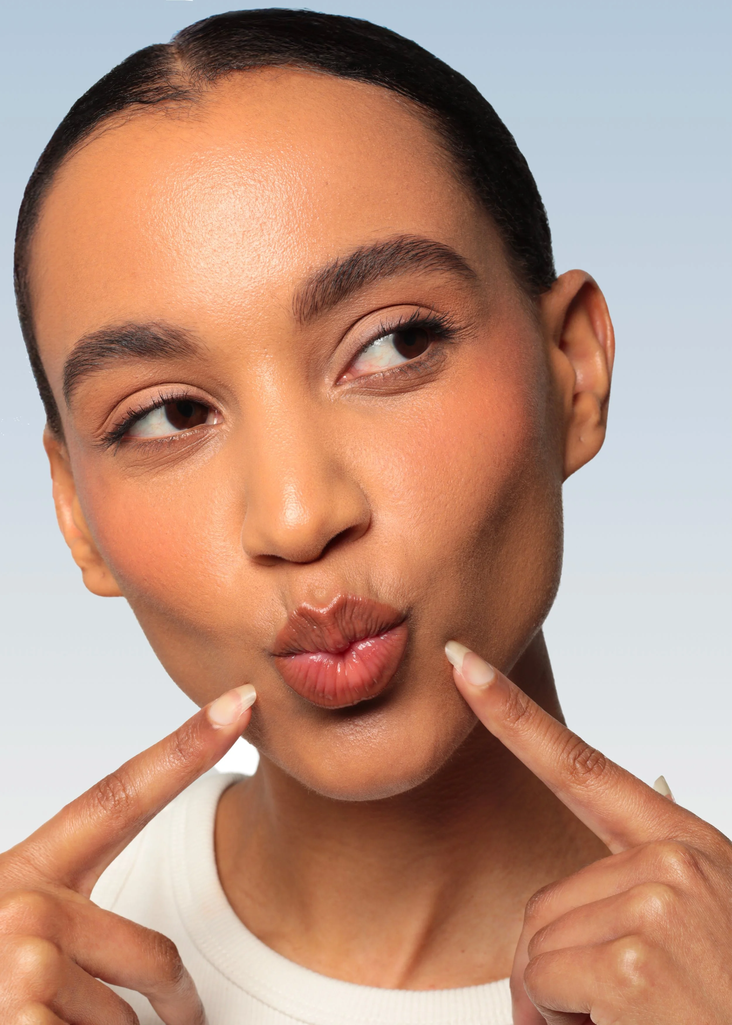 A young woman with dark hair, makeup, and clear skin making a playful expression by puckering her lips and pointing at her cheeks with both index fingers.