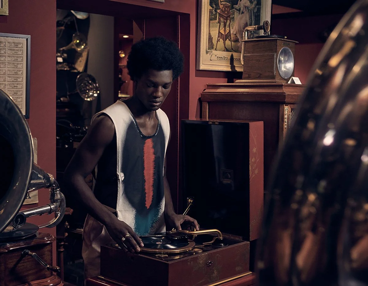 Person with dark skin and curly hair operating a vintage record player in a room with antique furniture and framed artwork on the wall.