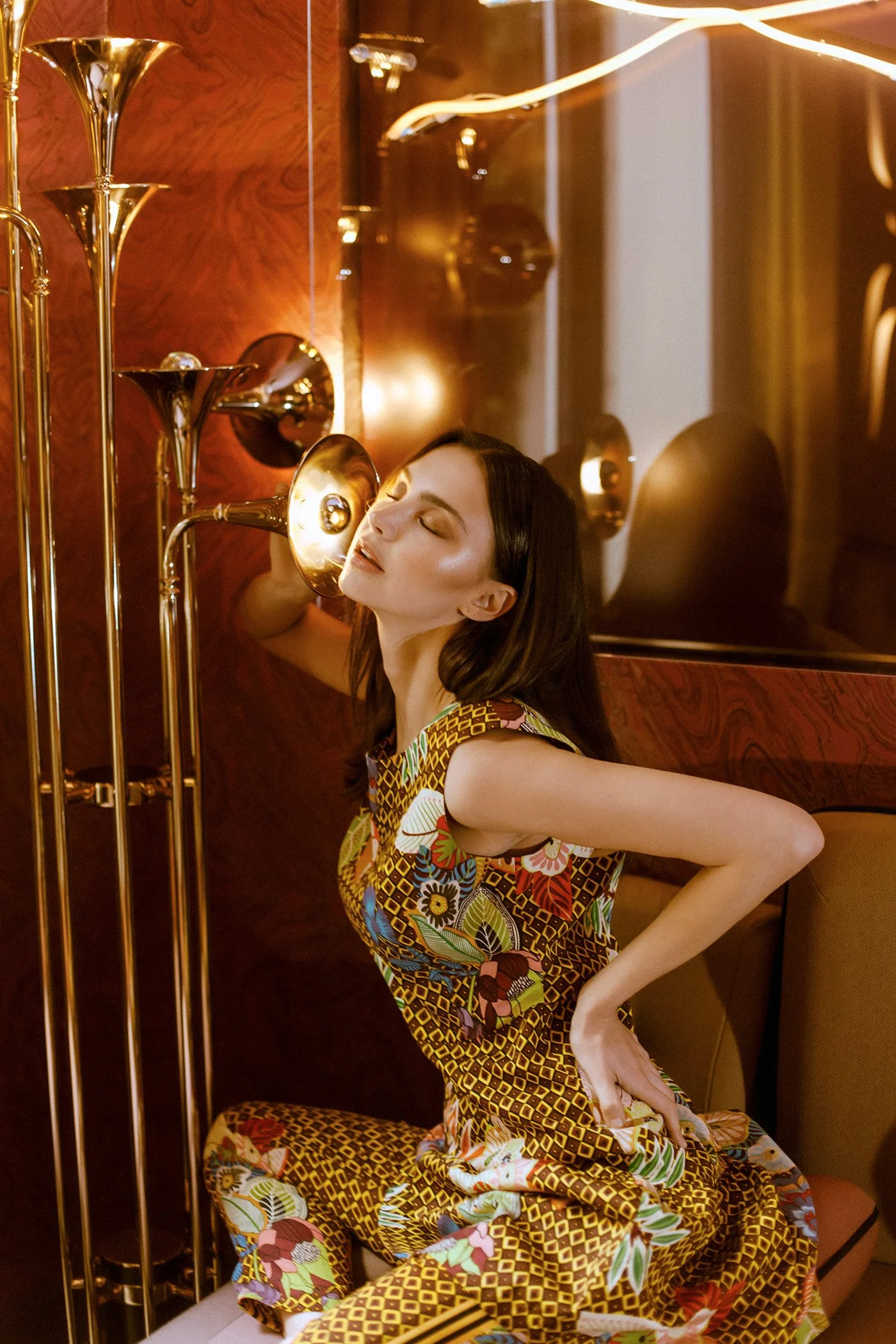 A woman with dark hair, dressed in a colorful floral patterned dress, is sitting on the floor in a room with warm, gold-toned lighting. She has her eyes closed, her head tilted back slightly, and one hand resting on her lower back, appearing to be in