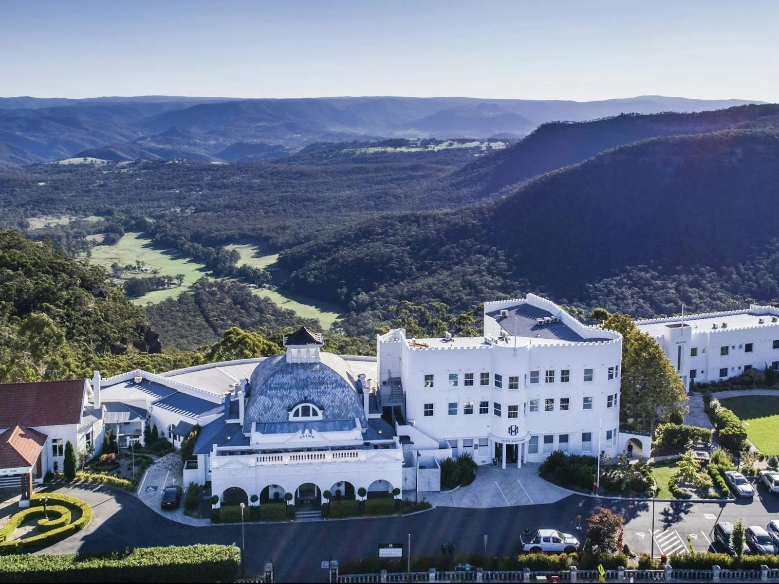 A large white building with a dome and turret, surrounded by parking lot with cars, set against green rolling hills and mountains in the background.