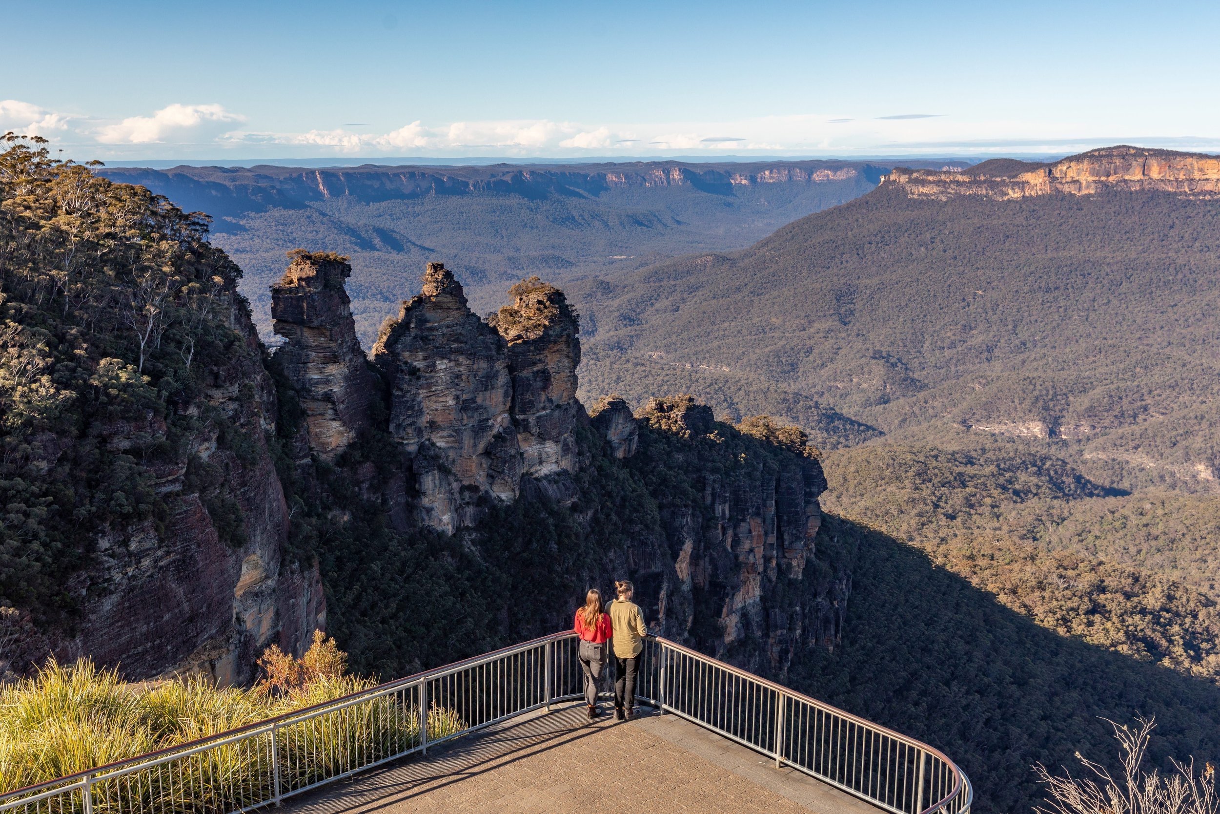 Two people standing on a viewing platform overlooking a vast canyon with rock formations, forested hills, and distant cliffs under a clear sky.