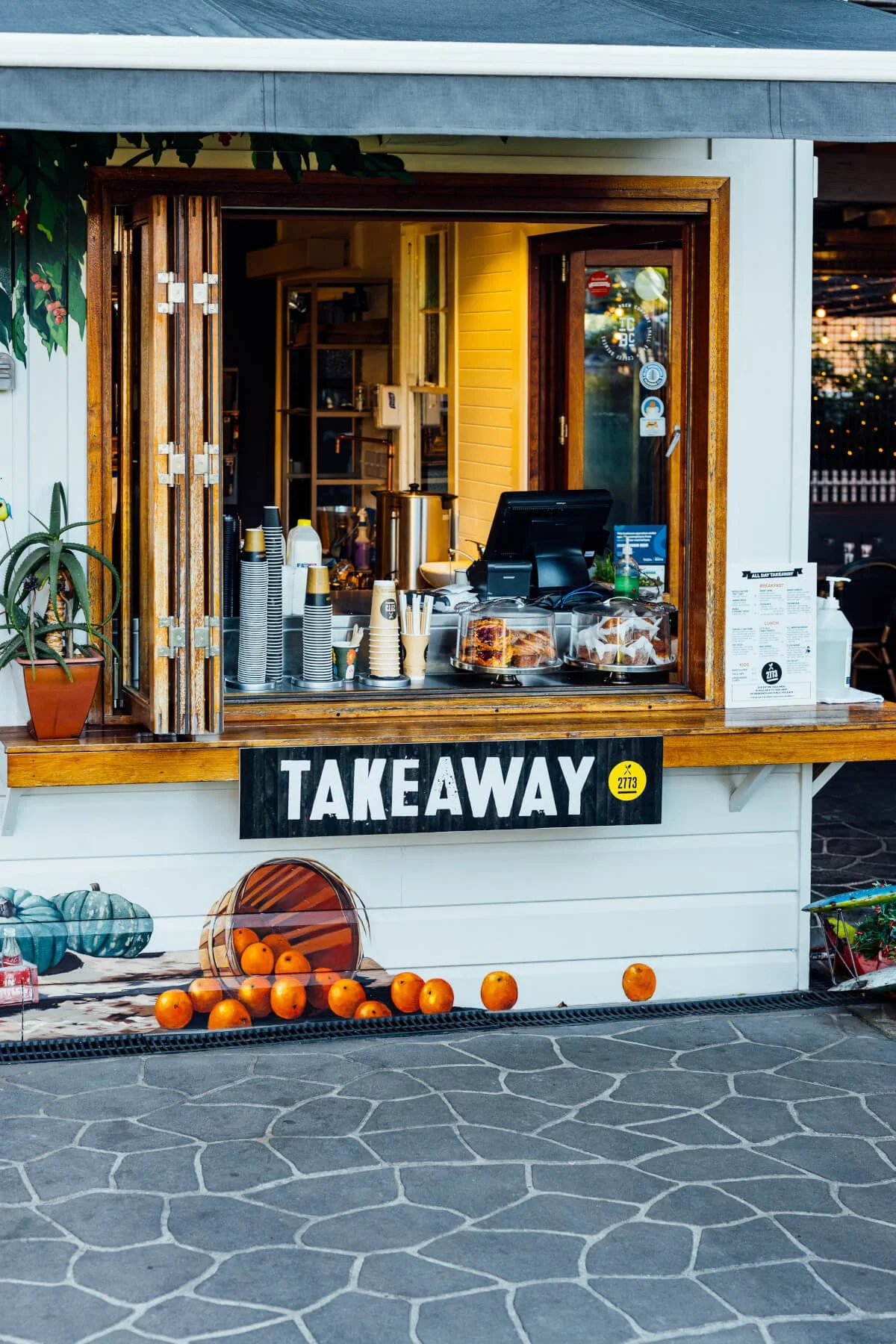 Outdoor takeout food stand with a black sign that says 'TAKEAWAY'. There are orange pumpkins with a bucket and a small white pumpkin at the base of the stand. Inside, cups, containers, and a cash register are visible.