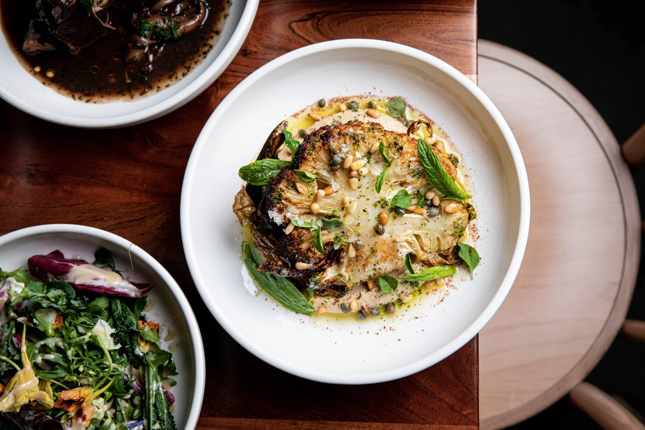 A roasted cauliflower steak garnished with pine nuts, basil, and green herbs on a white plate, with side salads visible in white bowls nearby.