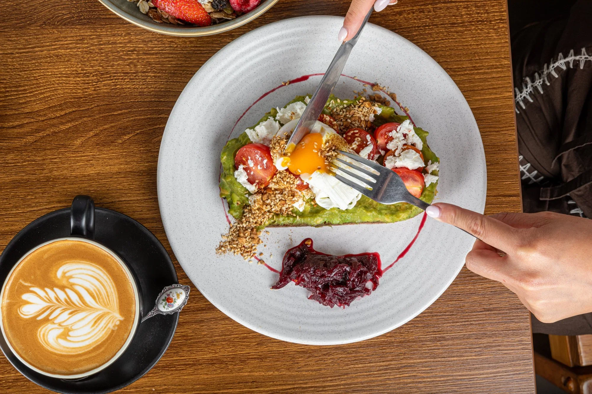 A white plate of avocado toast topped with cherry tomatoes, crumbled cheese, chopped nuts, a soft-boiled egg, and a drizzle of sauce, with a side of berry compote, on a wooden table. A cup of latte with latte art is beside the plate.