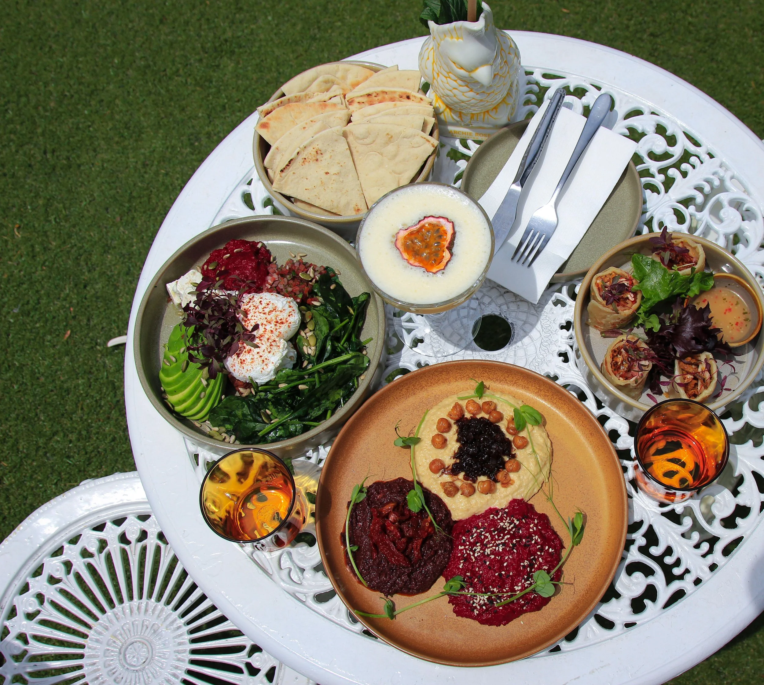 Outdoor table with various Middle Eastern dishes including hummus, tabbouleh, skewers, pita, and drinks.