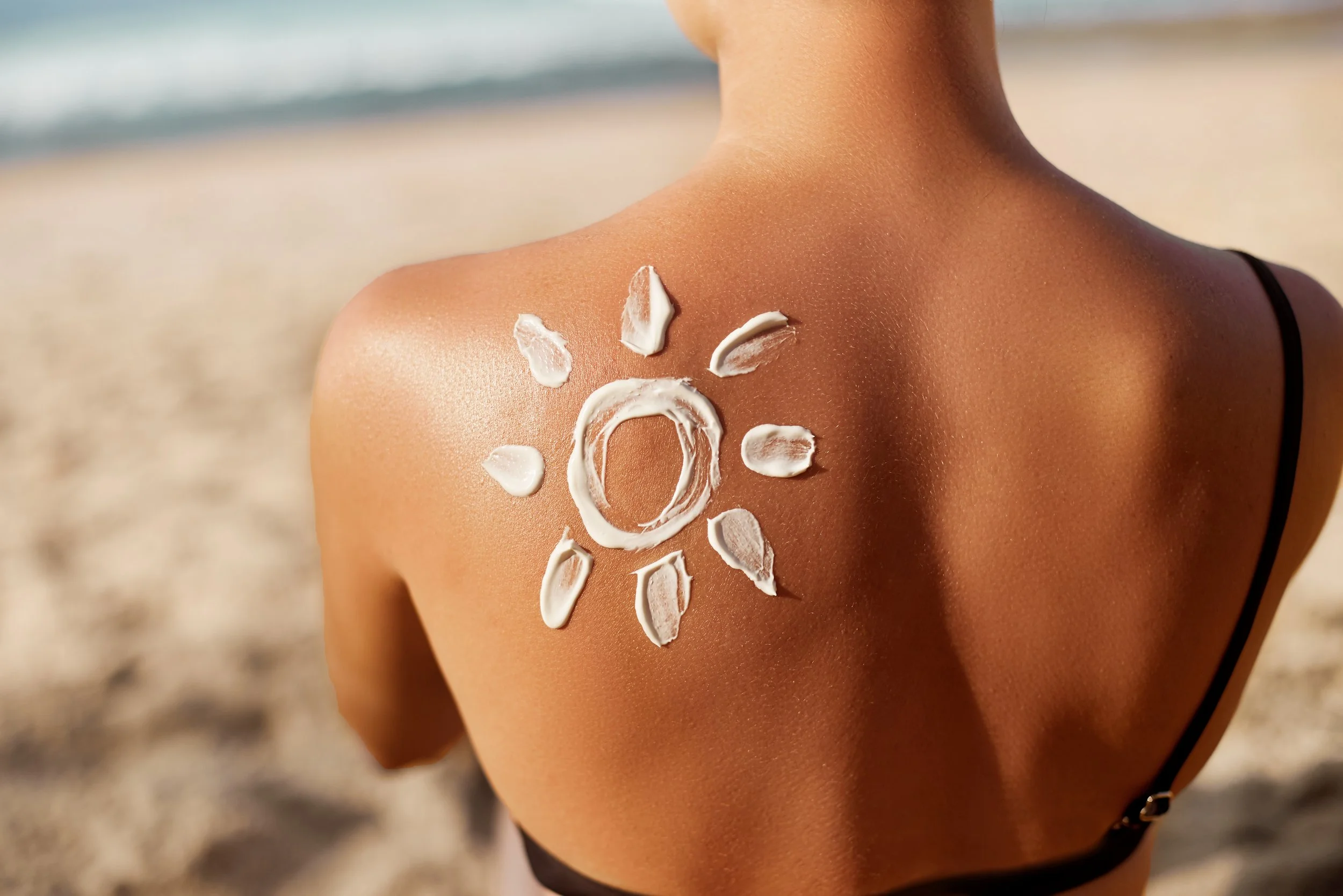 Back of a woman at the beach with sun lotion on her shoulder, shaped like a sun with the letter 'O' in the center.