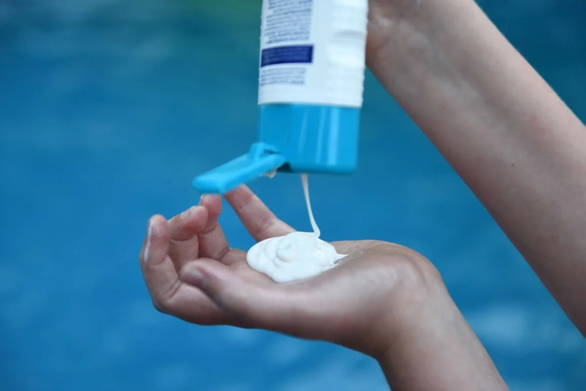 Person pouring white lotion or sunscreen from a blue and white bottle into their palm near a pool.