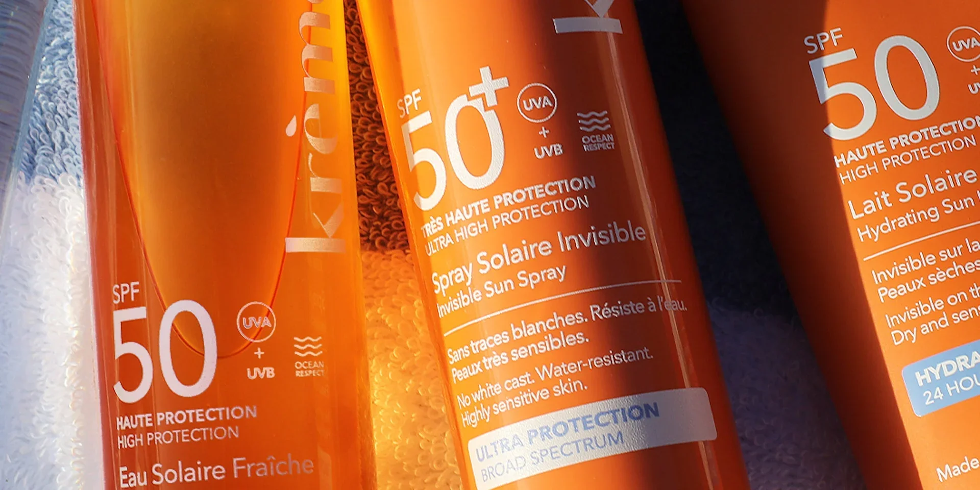 Three orange bottles of sunscreen with SPF 50, labeled as 'Invisible Sun Spray' and 'Hydrating Sun Lotion,' designed for high protection and suitable for sensitive skin, featuring UVA and UVB protection, water resistant, and broad spectrum.