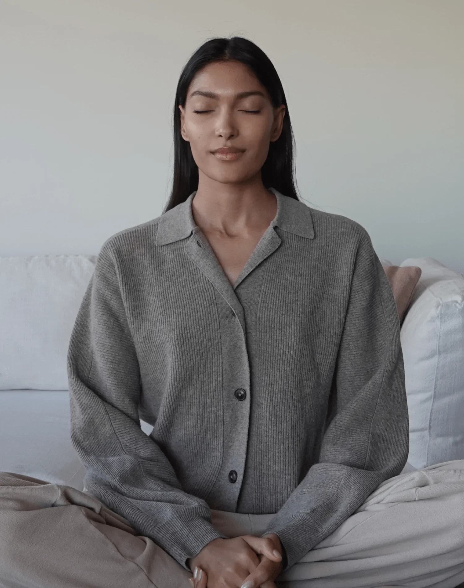 A woman sitting on a white sofa with eyes closed and a peaceful expression, wearing a gray button-up shirt.