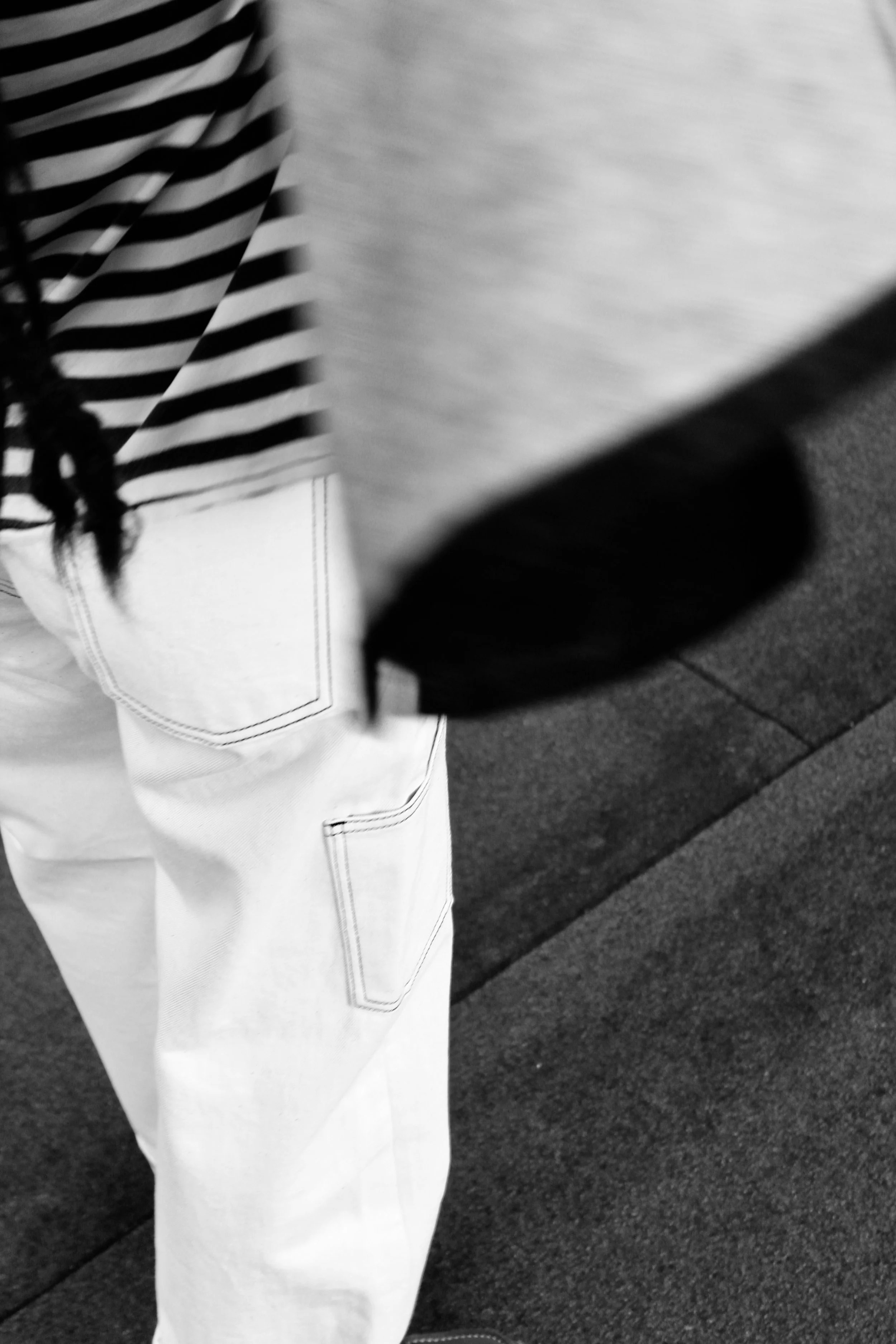 Partial view of a person wearing a striped shirt and white pants. The person is holding a book or notebook. The background appears to be a sidewalk or pavement.