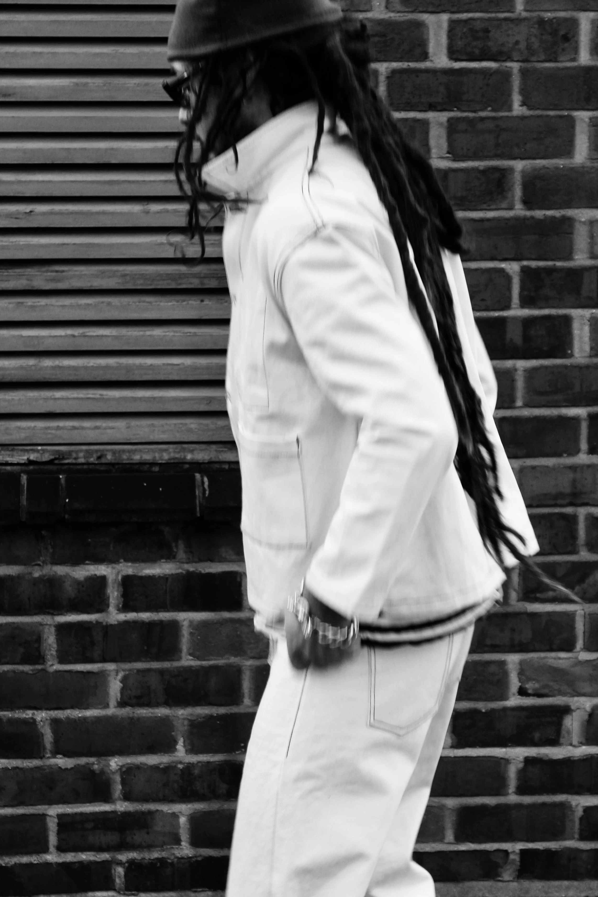 A person with long hair, wearing a beanie, glasses, a face mask, and a white jacket, standing in front of a brick wall and a wooden slatted background.