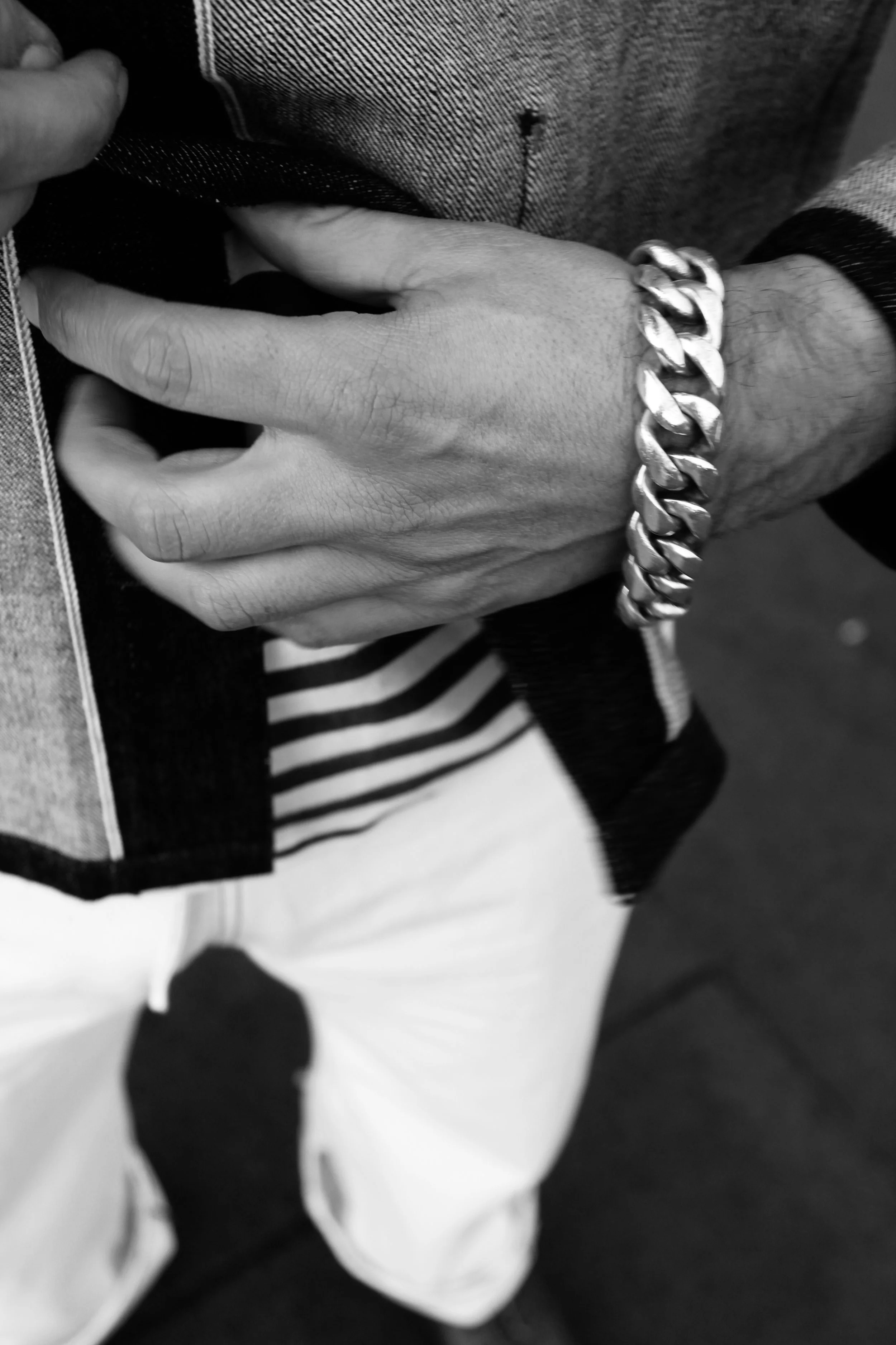 Close-up of a person's hand with a chunky chain bracelet. The person wears a long-sleeve jacket and striped pants, and the background is blurred.
