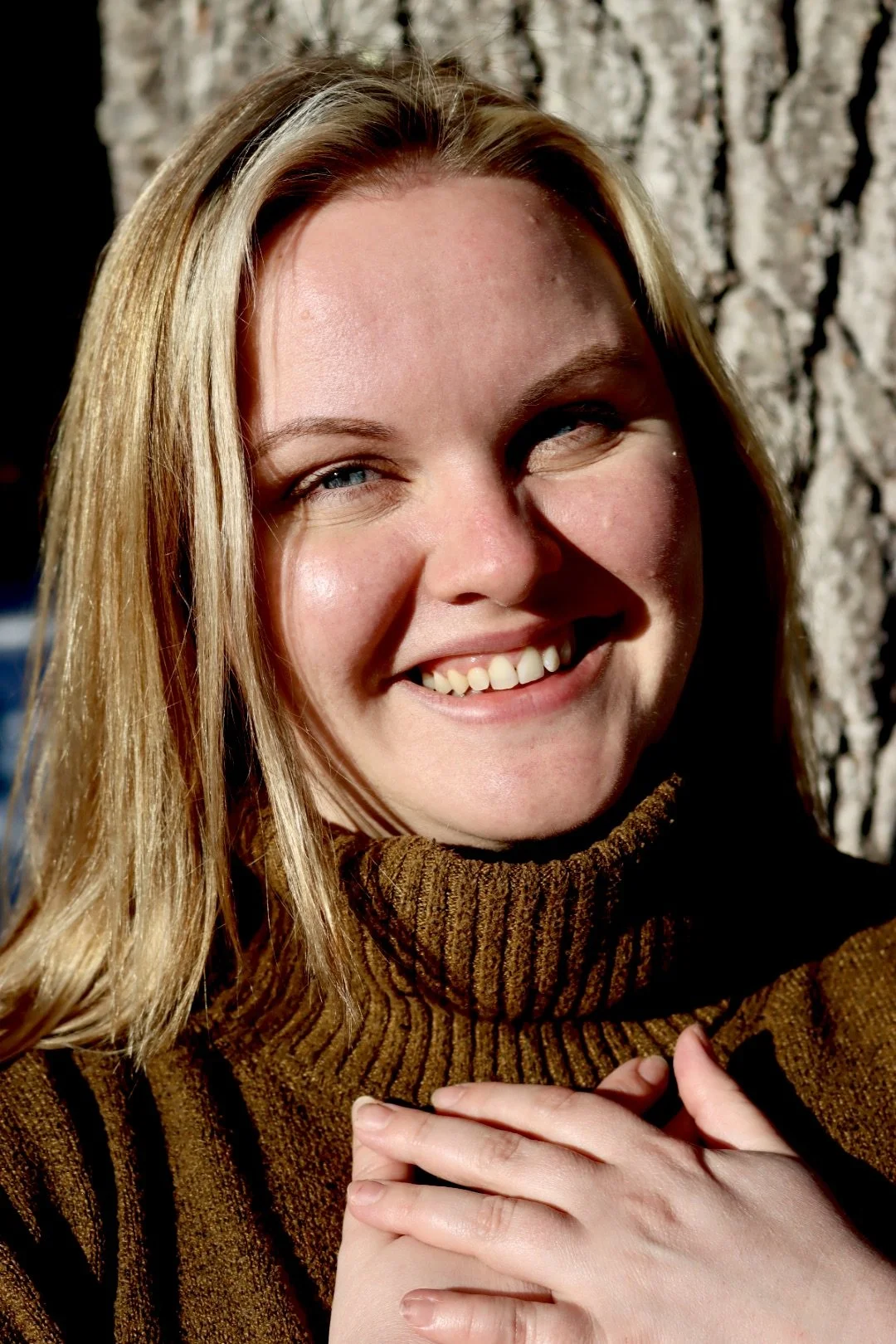 Close-up of a smiling woman wearing a brown turtleneck sweater, outdoors with a tree trunk in the background.