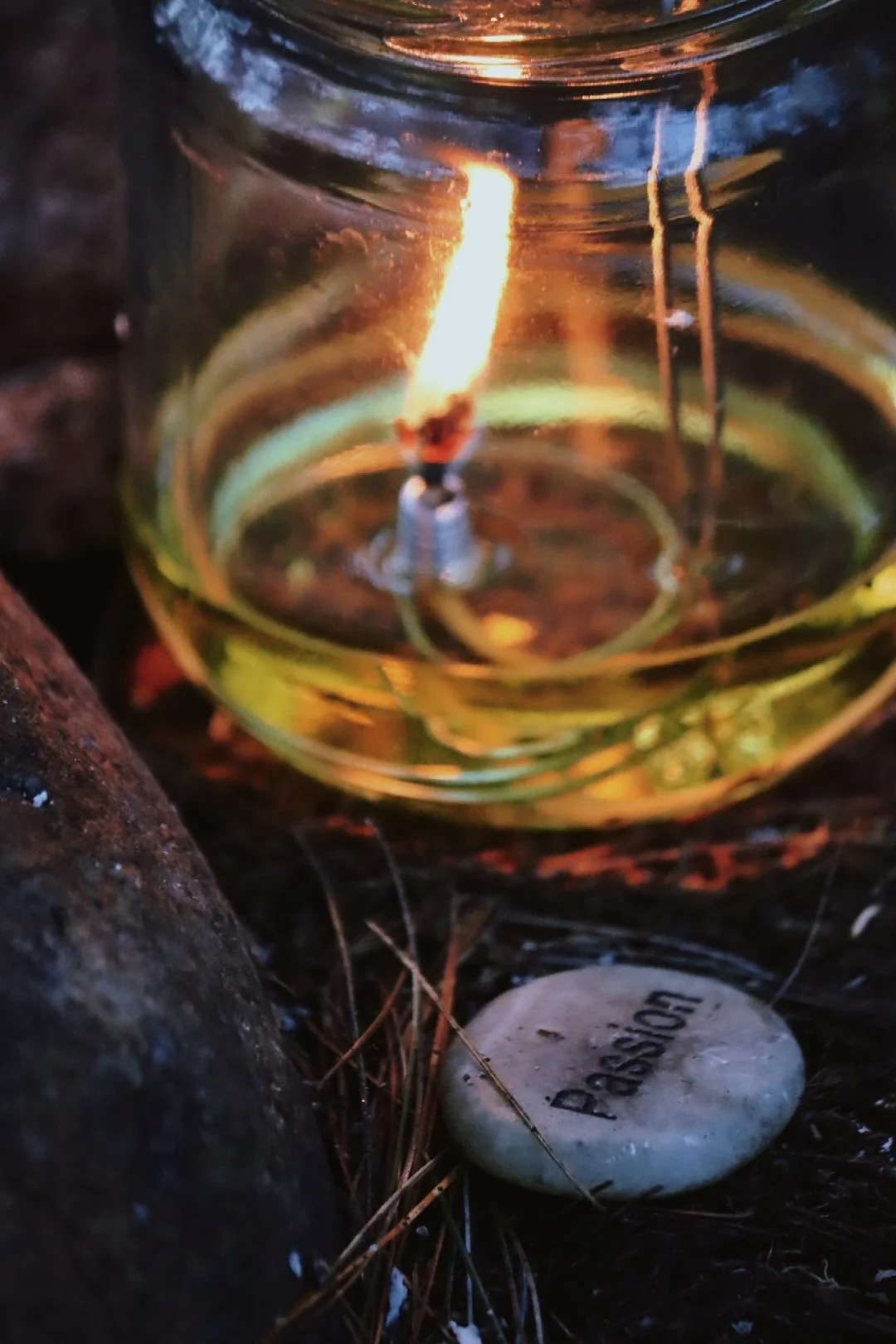 Close-up photograph of a lit candle inside a glass container, placed on the ground among rocks and pine needles, with a nearby white stone engraved with the word 'Pray'.