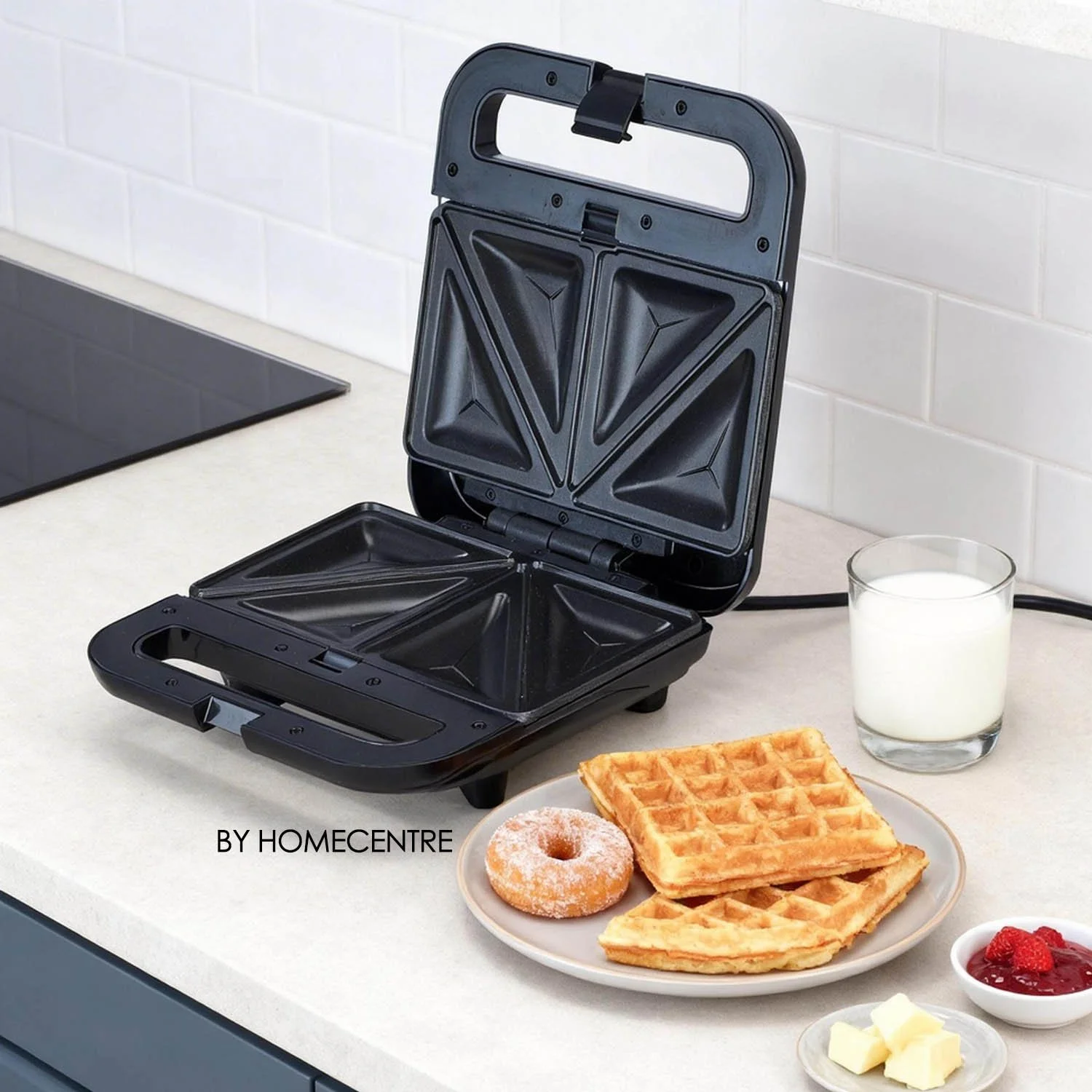 Black waffle maker open on a kitchen countertop with a plate of waffles, a donut, a glass of milk, butter, and jam nearby.