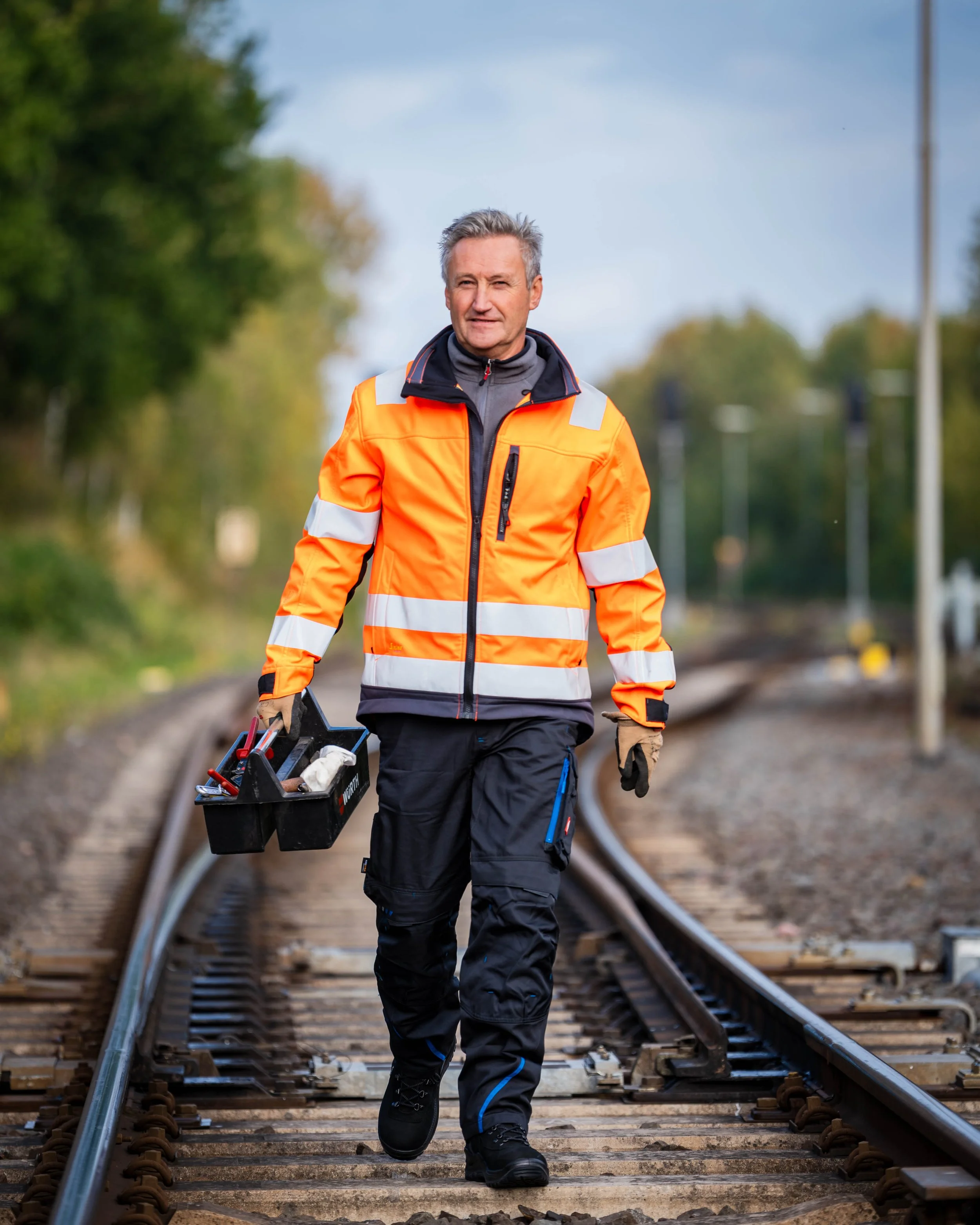 Ein Mann in einer orangefarbenen Warnjacke läuft auf Gleisen und trägt eine Werkzeugkiste.