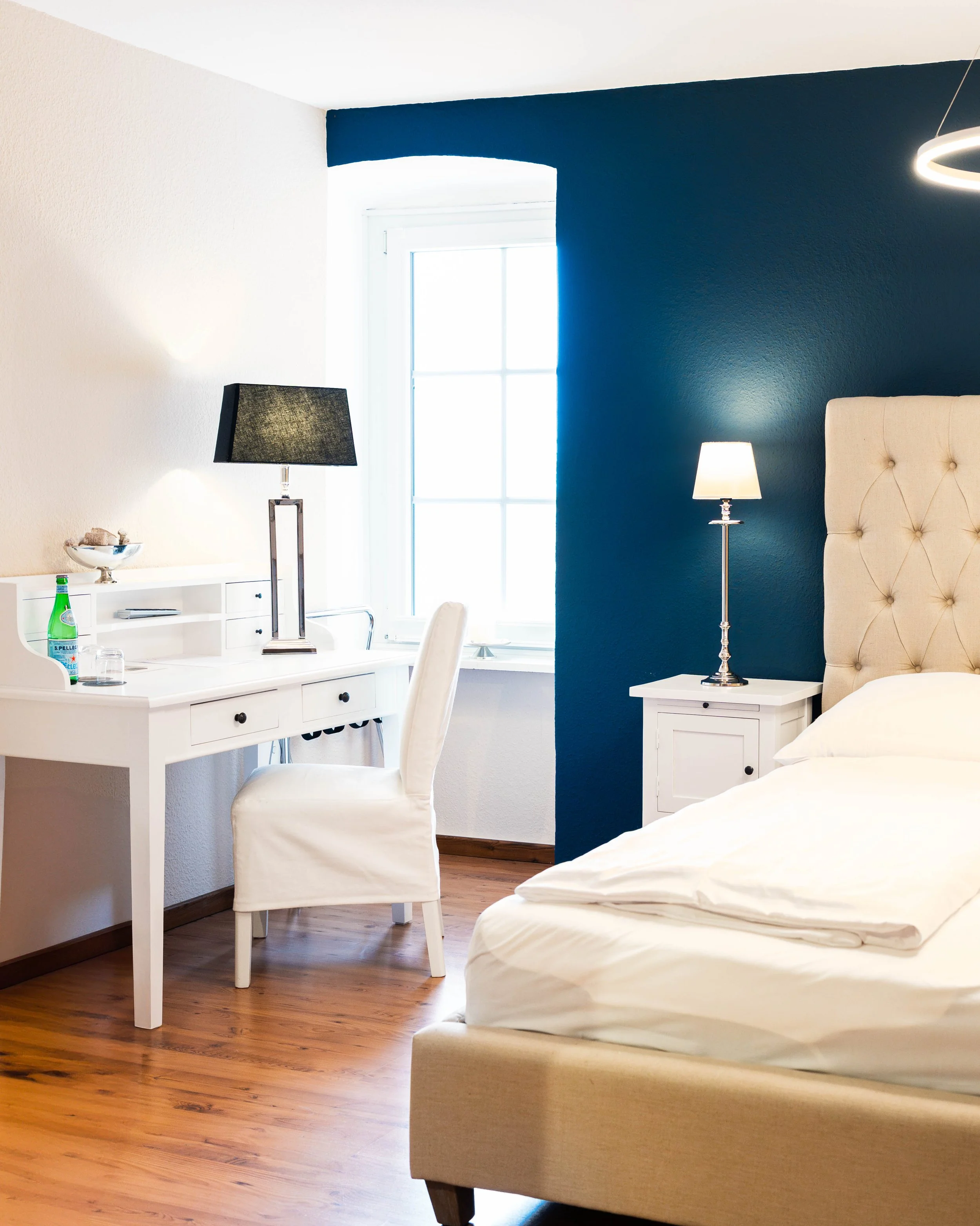 Ein Schlafzimmer mit weißem Bett, einem weißen Nachttisch mit Stehlampe, einem weißen Schreibtisch mit dunklem Lampenschirm, einer weißen Stuhllehne, einer blauen Akzentwand und einem Fenster.