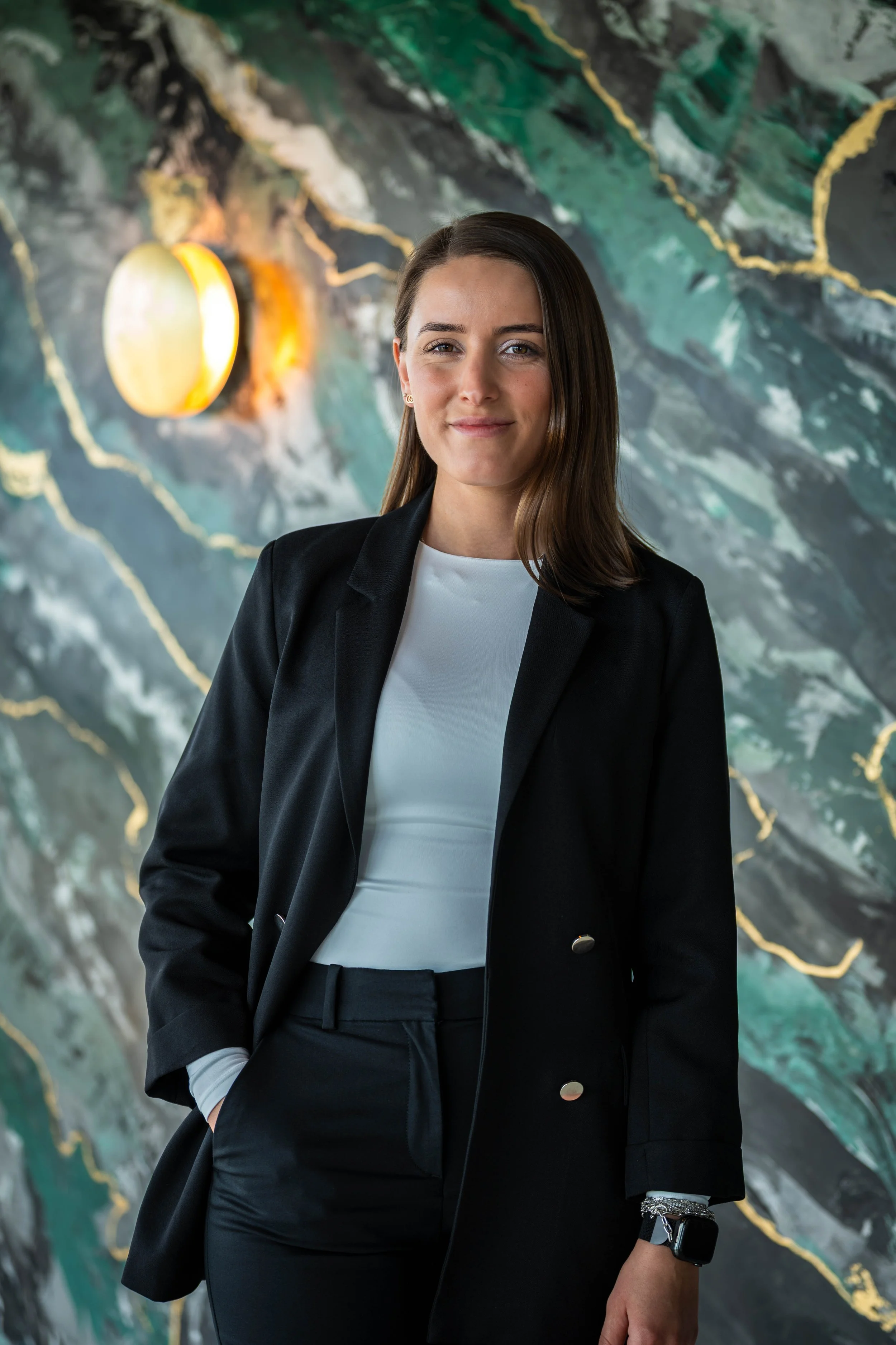 Eine junge Frau in einem schwarzen Anzug und weißem Oberteil steht vor einer künstlerischen Wand mit grünen, weißen und schwarzen Mustern und einem beleuchteten runden Element im Hintergrund.