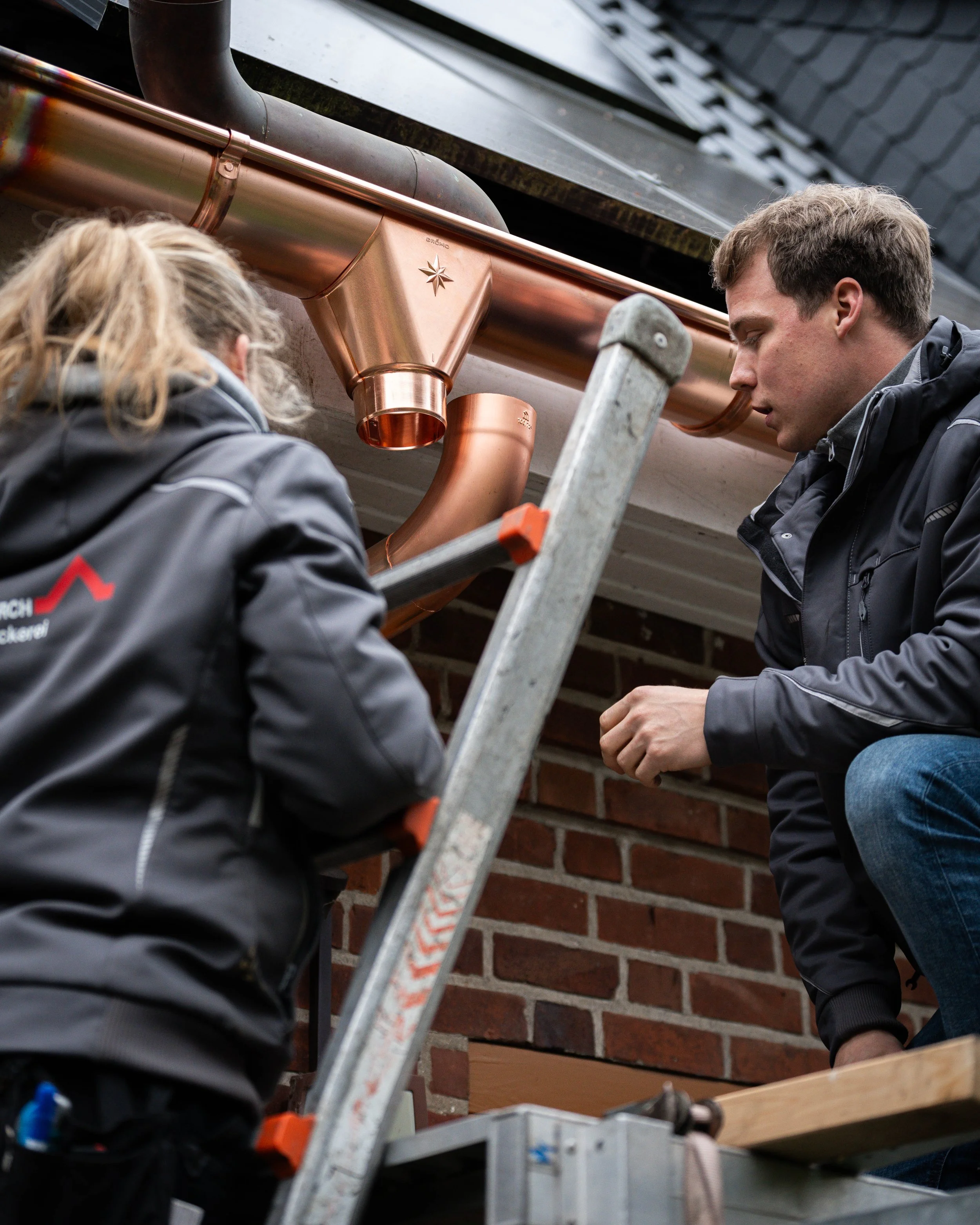 Zwei Handwerker arbeiten an einem Kupferdach, eine Person steht auf einer Leiter, eine andere ist kniend.