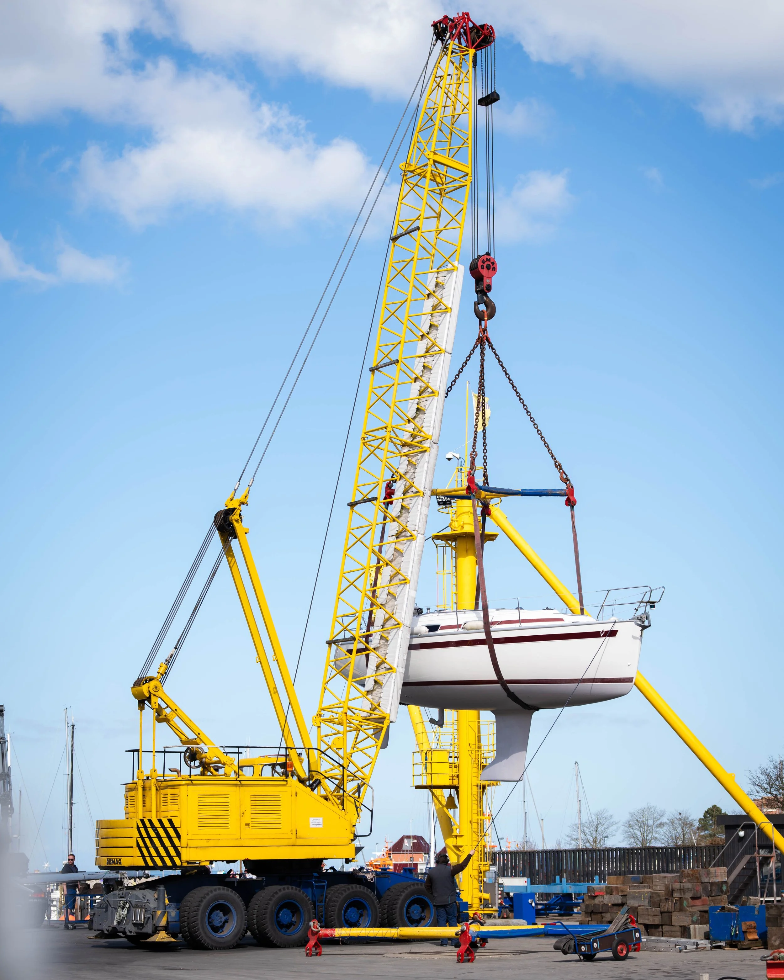 Ein Kran hebt ein Segelboot über den Boden. Der Kran ist gelb, die Haken sind rot, und das Segelboot ist weiß mit einem schwarzen Streifen. Es ist ein sonniger Tag, blauer Himmel mit Wolken, und es gibt einige Leute im Hintergrund.