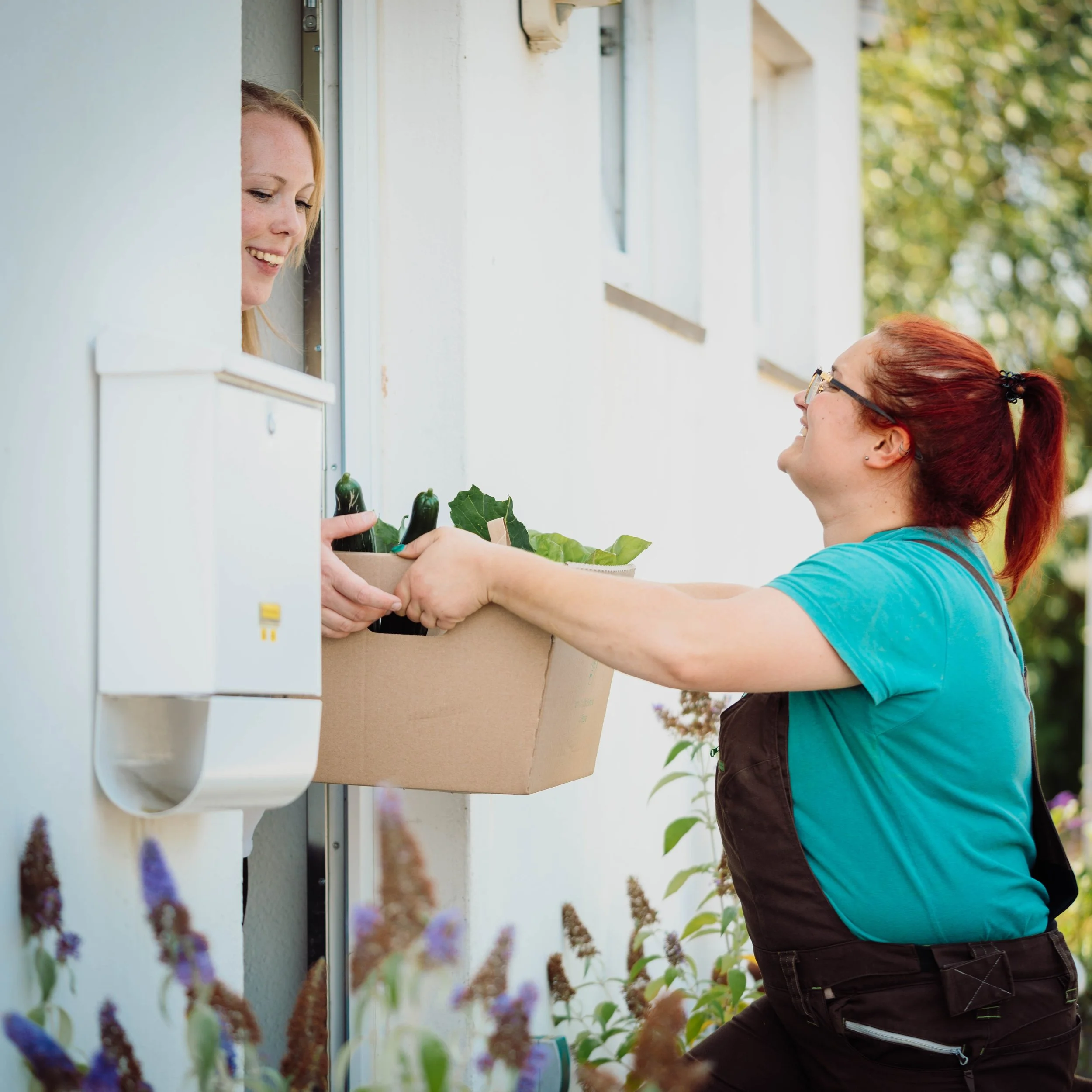Zwei Frauen tauschen Gemüse an einer Haustür in einem Garten aus.