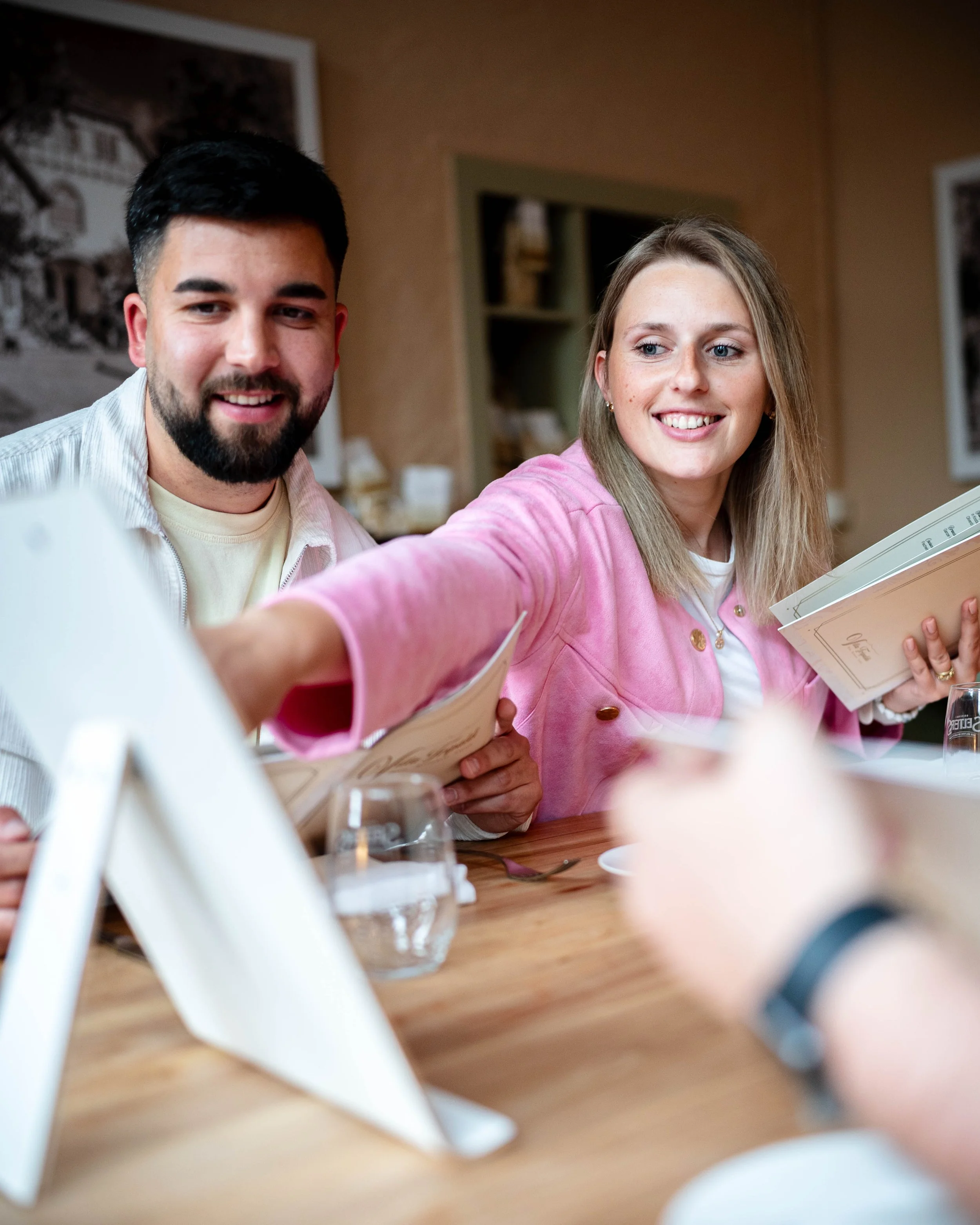Zwei lachende Menschen, ein Mann mit dunklem Haar und Bart, eine Frau mit blonden Haaren, sitzen an einem Tisch in einem Restaurant, während die Frau die Kamera oder den Betrachter anspricht.