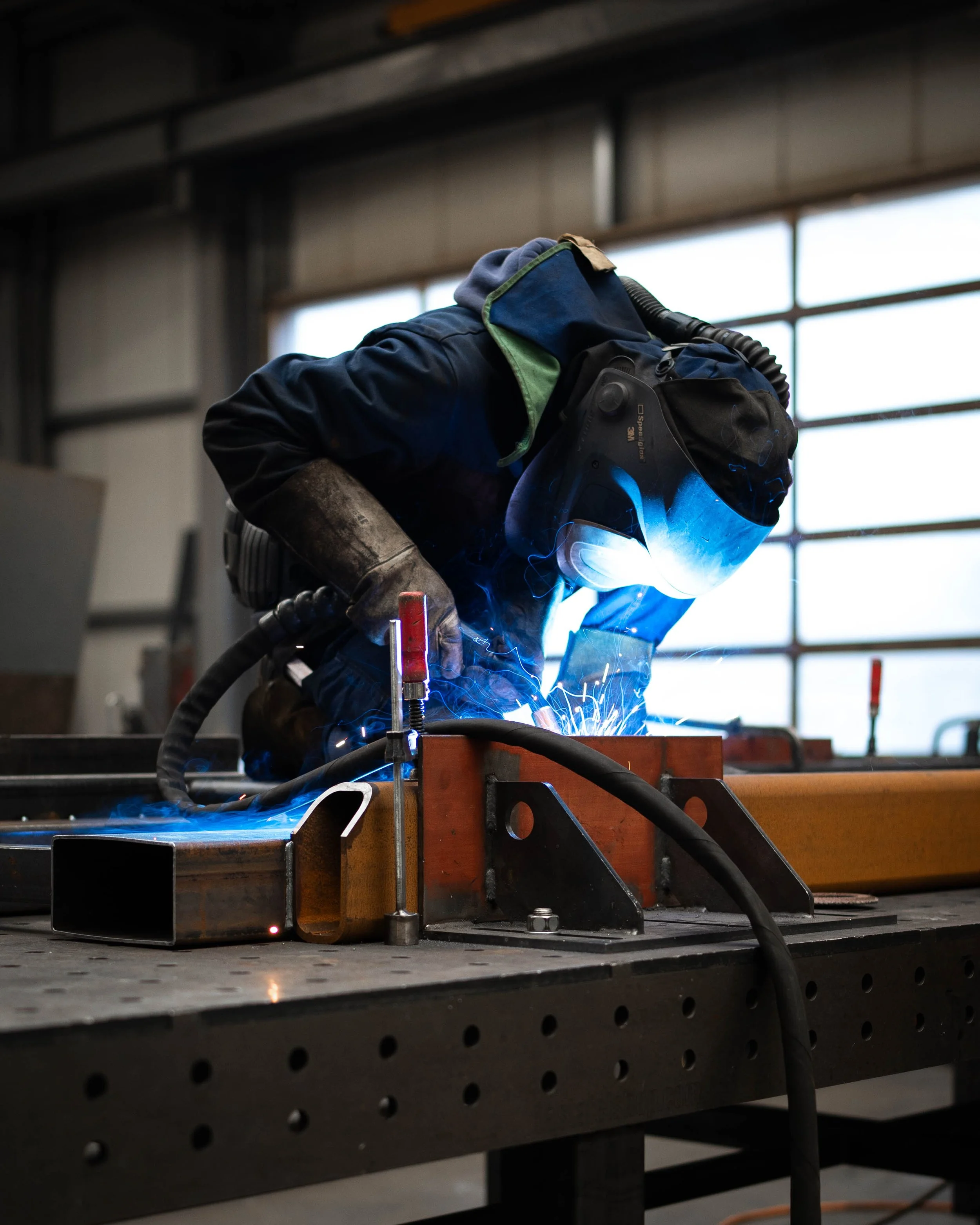 Schweißer in Schutzkleidung arbeitet an Metallstück in Werkstatt mit Flammen und Funken.
