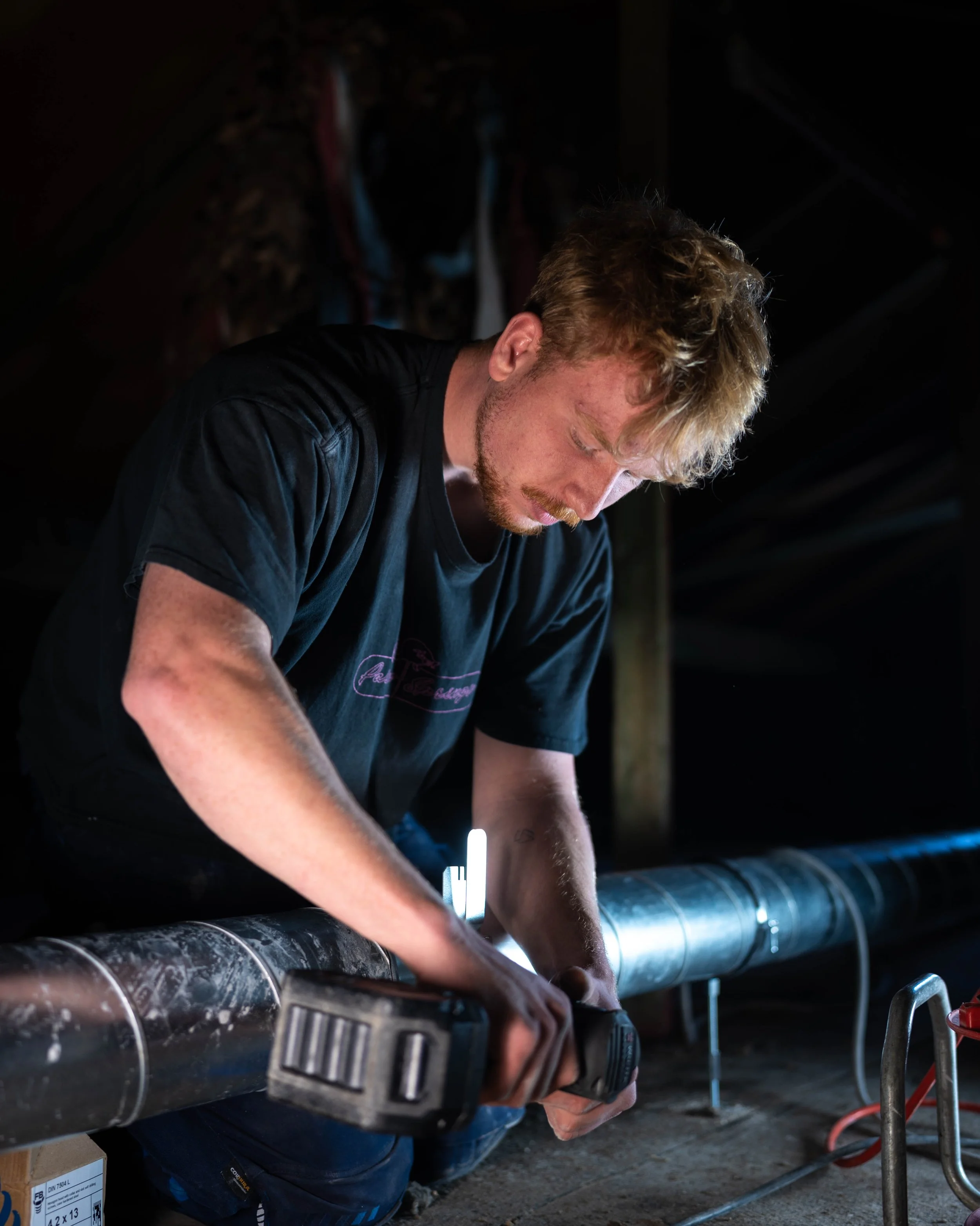 Ein junger Mann arbeitet mit einem Akku-Schrauber an einer großen Rohrleitung in einem dunklen Raum.
