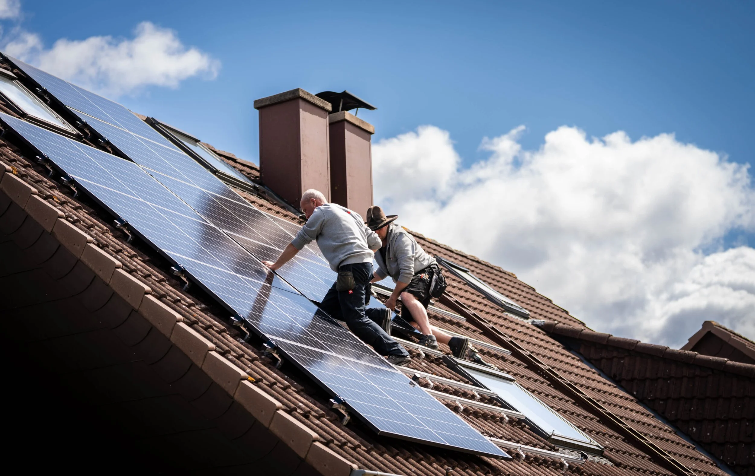 Zwei Menschen installieren Solarpaneele auf einem neungeschossigen Dach mit roten Ziegeln bei sonnigem Himmel.