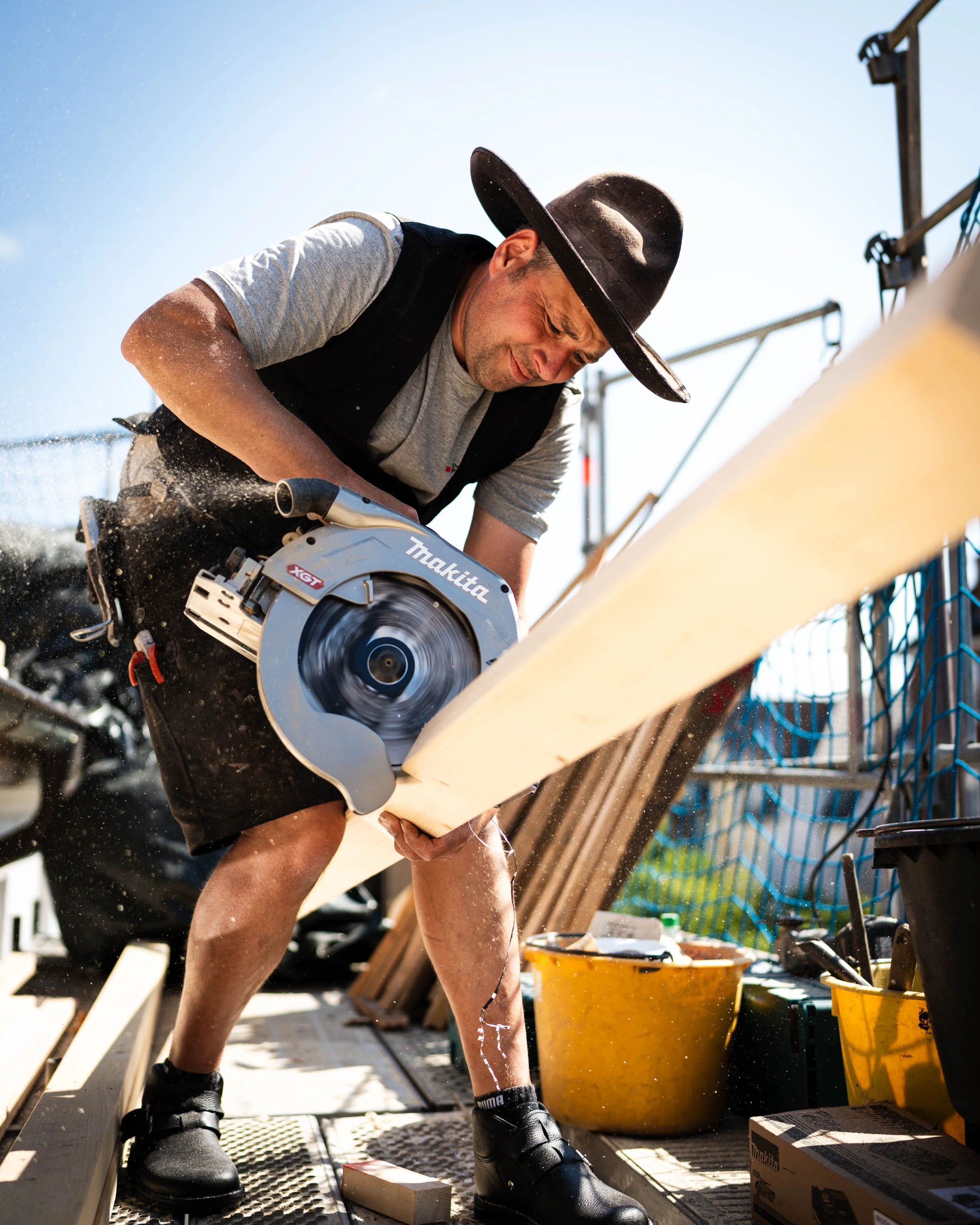 Ein Mann, der mit einer Kreissäge Holz schneidet, trägt einen Hut und sitzt auf einem Holzbohlen auf einem Baugerüst.