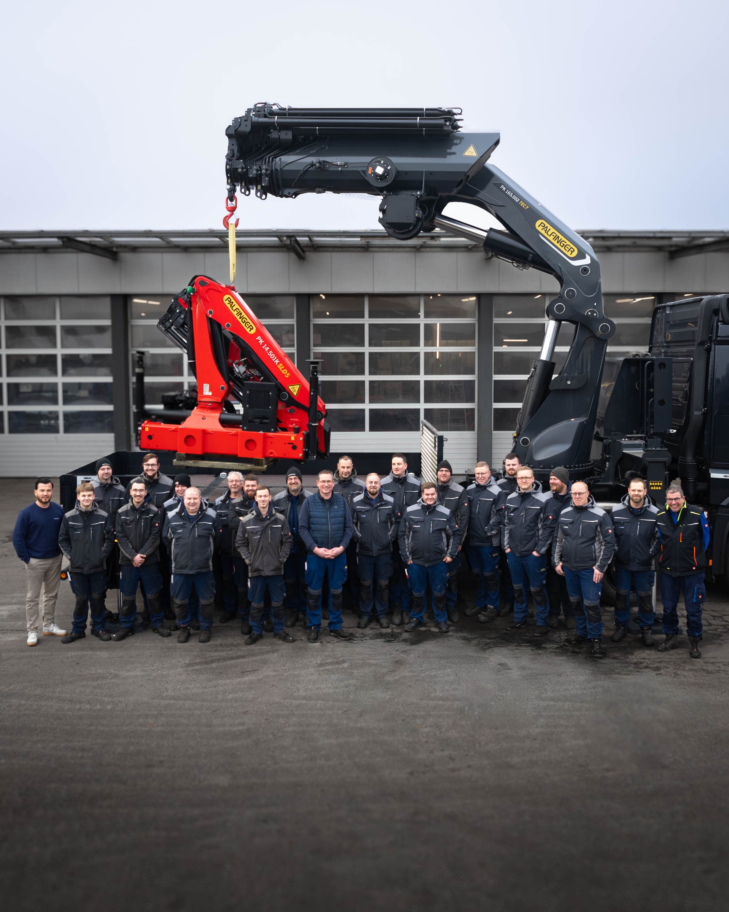 Eine Gruppe von Menschen steht vor großen Hebezeugen, darunter ein rotes und ein schwarzes technisches Gerät, vor einer modern wirkenden Gebäudefassade mit großen Fensterflächen.