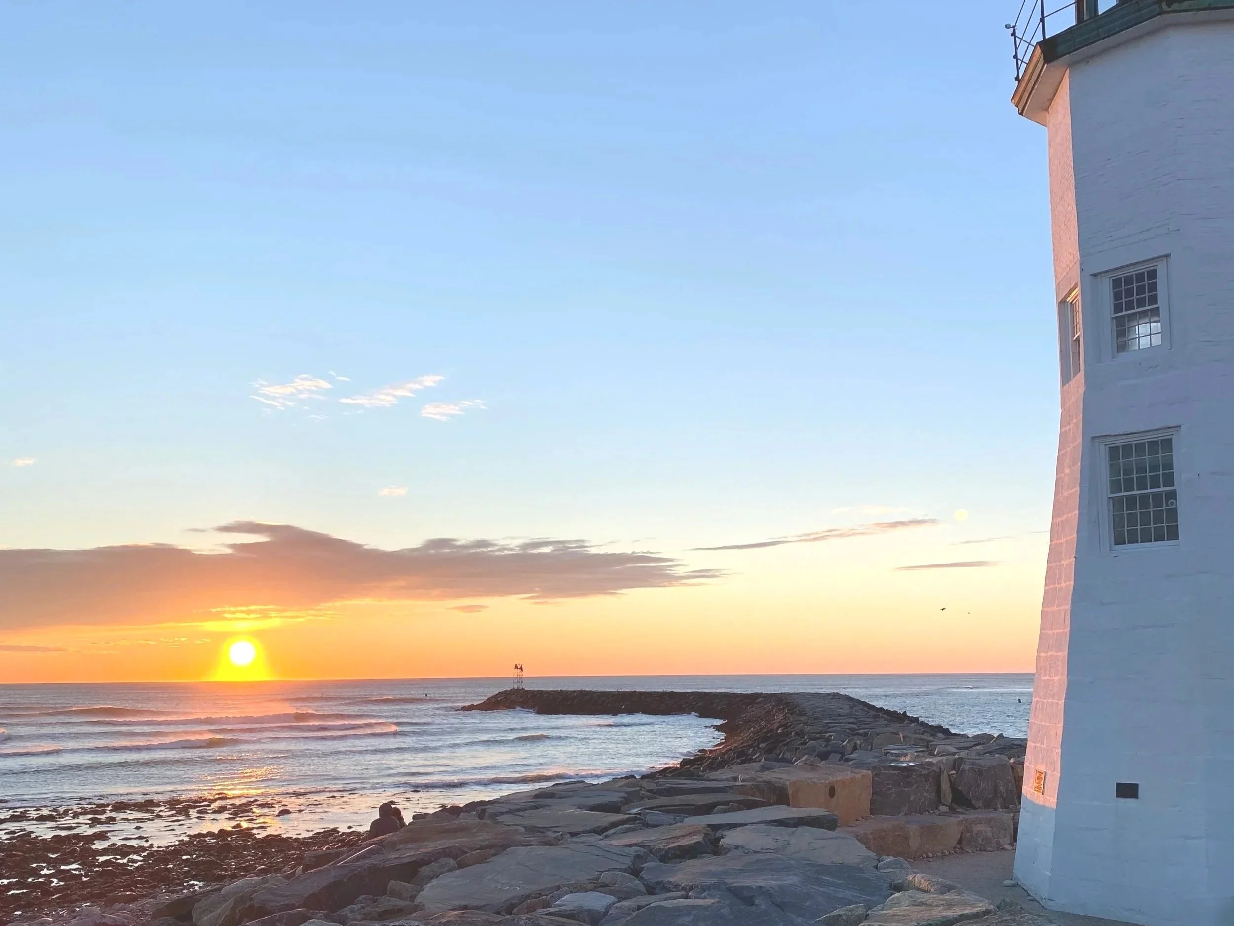 Sunset over the ocean with a lighthouse on a rocky breakwater, and a white building to the right under a clear sky.