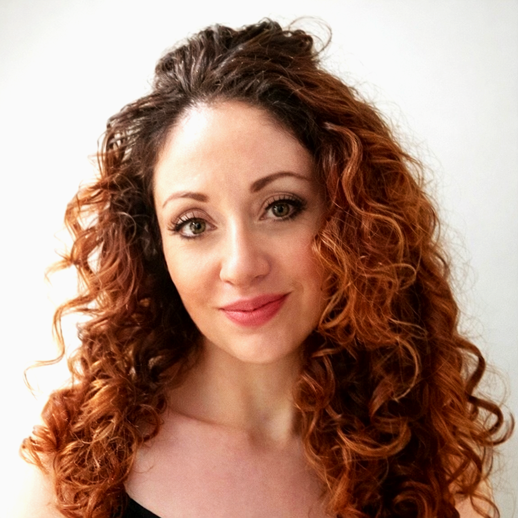 Portrait of a woman with curly red hair, light skin, and a gentle smile, wearing a light-colored top, against a plain white background.