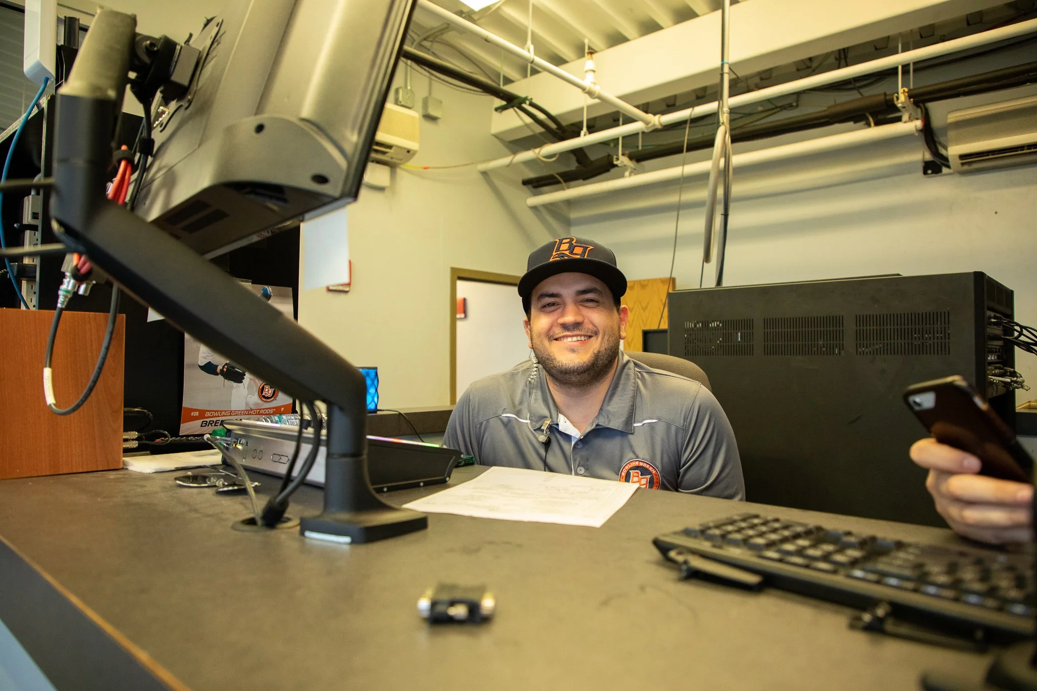 A young man wearing a black baseball cap and gray polo shirt sitting at a desk with a computer monitor, keyboard, and remote control, smiling and holding a smartphone in his right hand, in an office or control room setting.