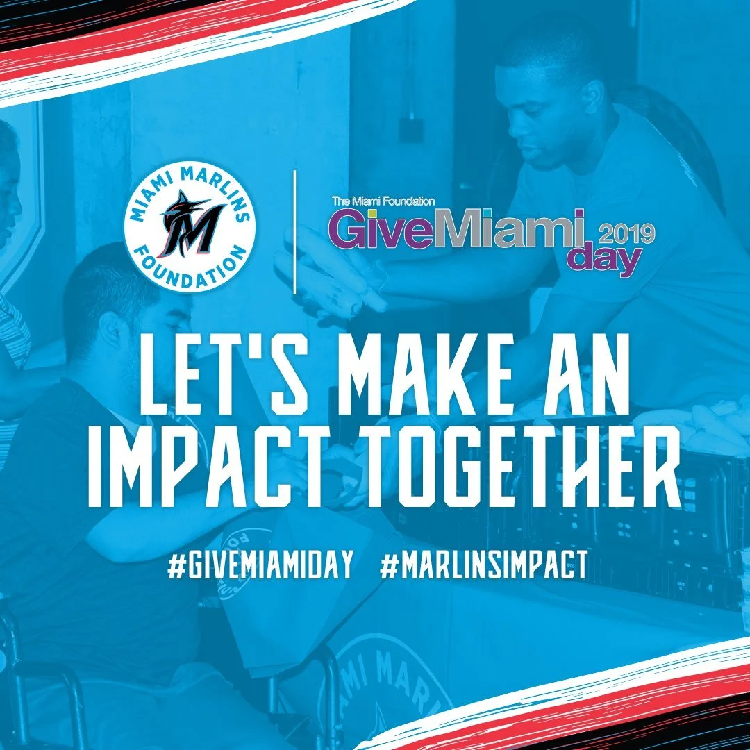 Group of people at a table with a promotional sign for Miami Marlins Foundation and GiveMiami Day 2019. The text says, 'Let's make an impact together' with hashtags #GiveMiamiDay and #MarlinsImpact.