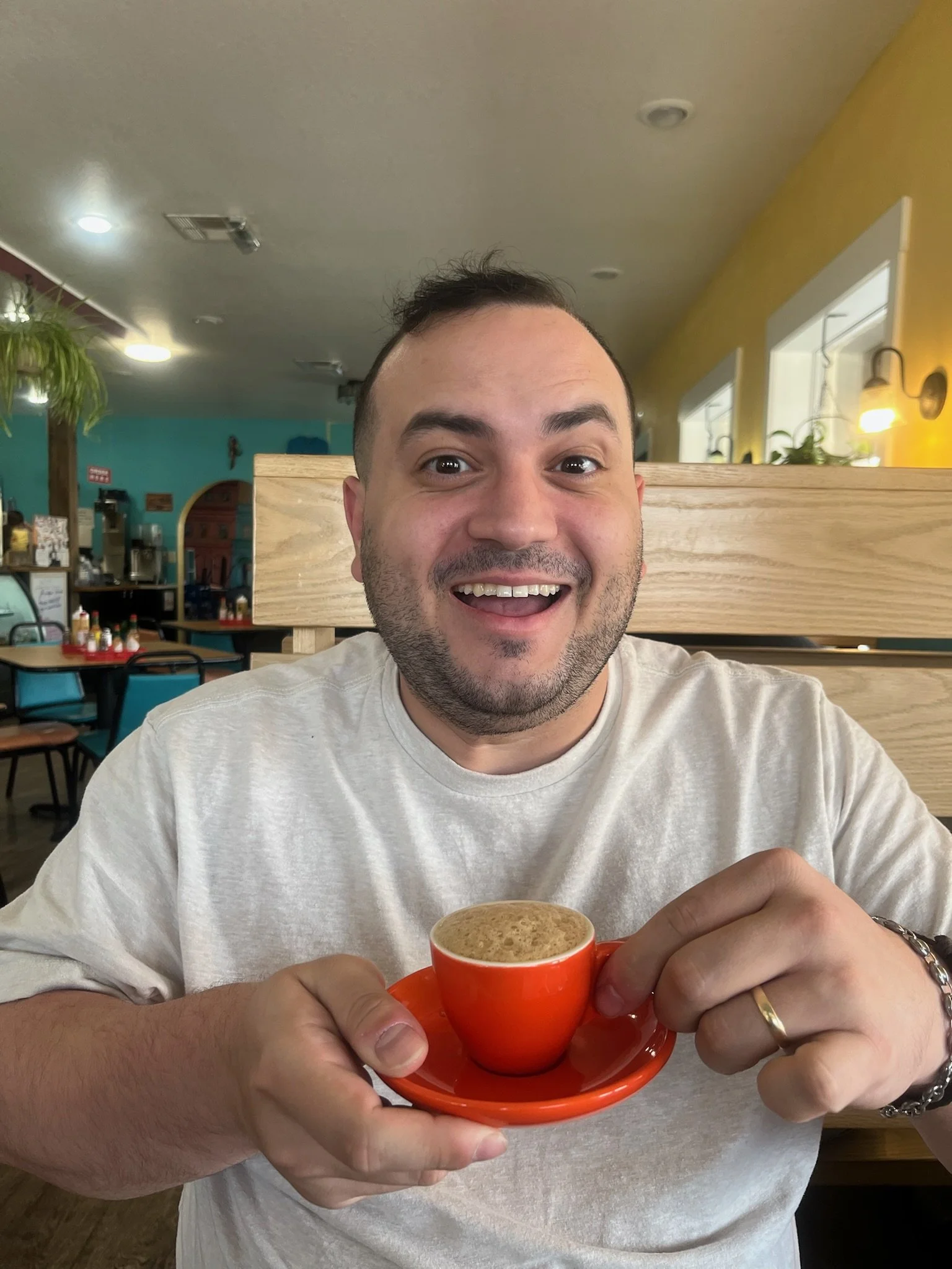 A man with a big smile holding a small orange cup of coffee in a cafe.