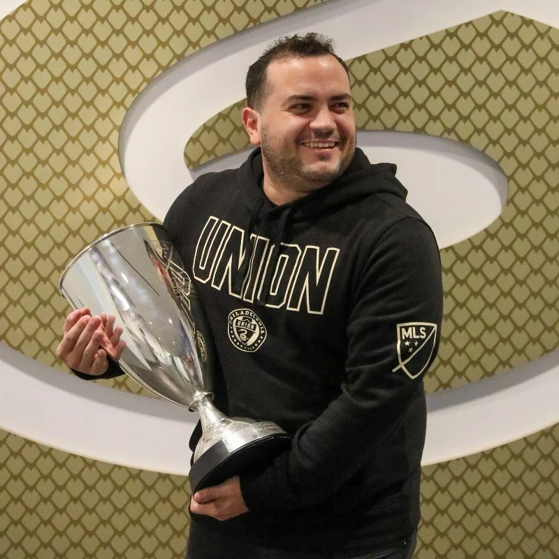 A man smiling and holding a large silver trophy in front of a patterned gold and white background.