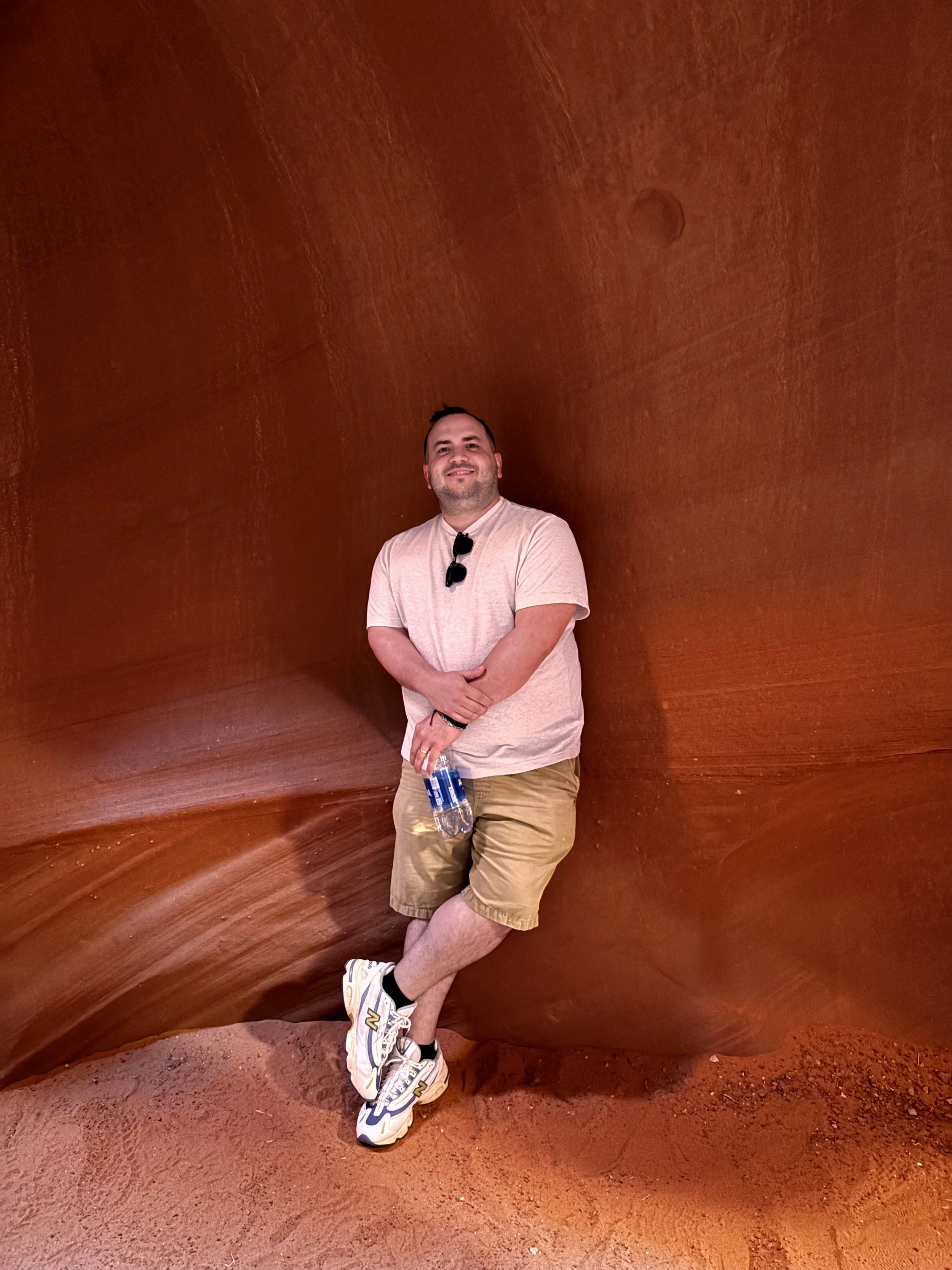 A man wearing a light pink t-shirt, beige shorts, and sneakers is posing inside a large, smooth, reddish-brown rock formation with swirling patterns. He is holding a water bottle in his right hand, has sunglasses hanging on his shirt, and is standing with one foot crossed over the other, smiling at the camera.