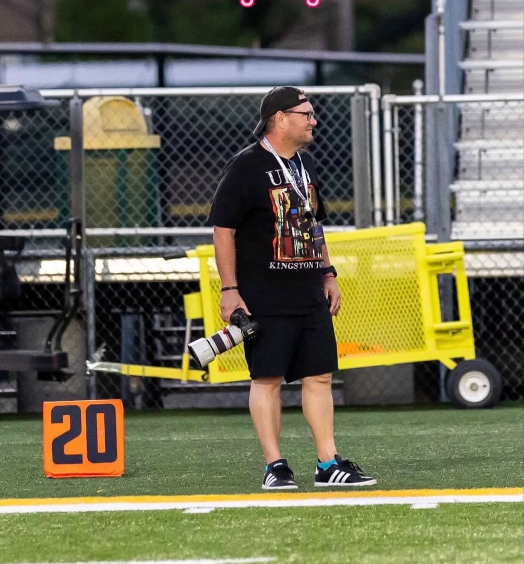 A man standing on a football field with a camera in hand, wearing a black cap, black T-shirt, black shorts, and athletic shoes, next to an orange yard marker with the number 20.