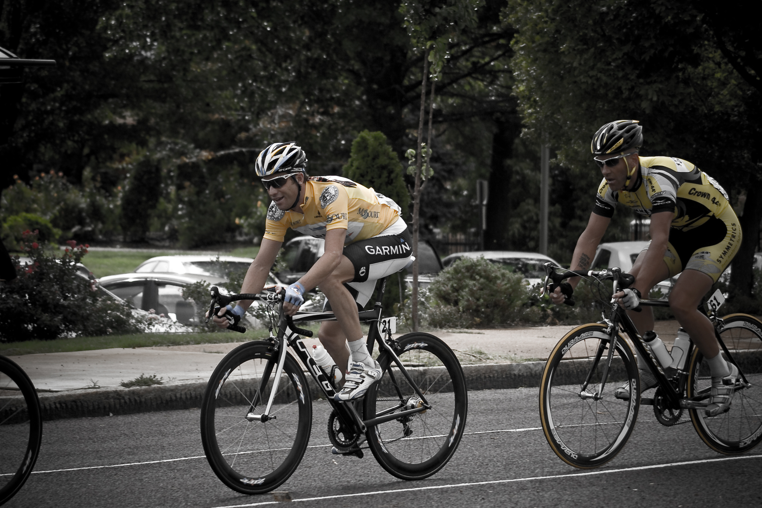 Cyclists in a race, wearing yellow and black jerseys, riding on a road with trees and parked cars in the background.