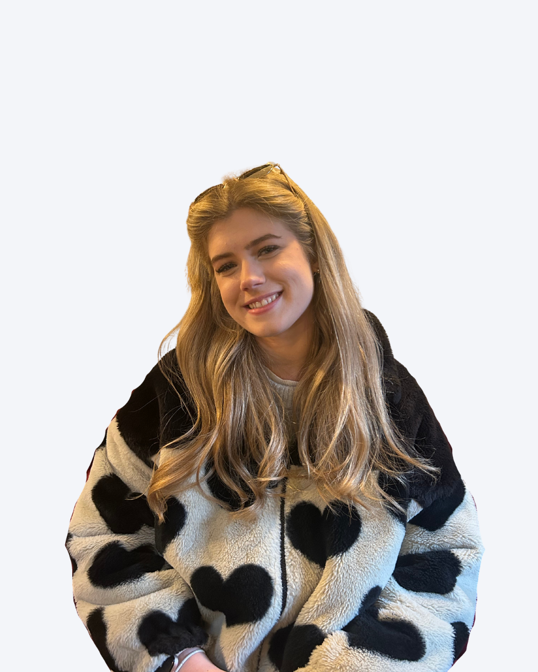 Young woman with long blonde hair smiling, wearing a black and white fleece jacket with heart patterns, sitting against a plain light background.
