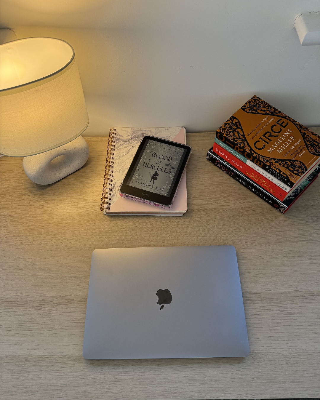 A desk with a closed silver Apple MacBook, a gray lamp, a pink notebook, a Kindle displaying the book 'Blood of Hercules', and a stack of books including 'Circe' by Madeline Miller, 'Fairy Tales' by Mairi Maclennan, and others.
