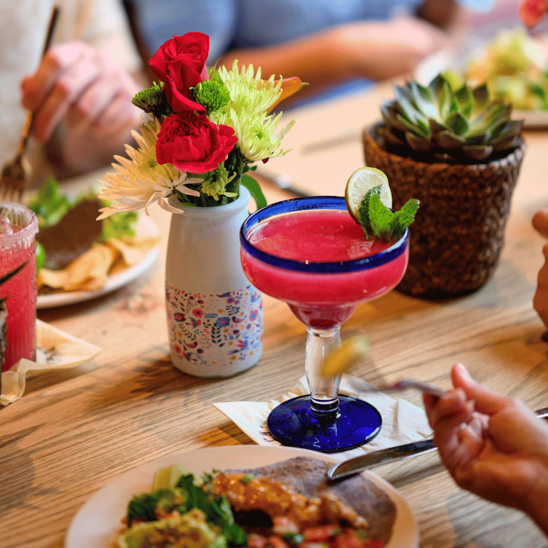 A colorful pink cocktail with a lime wedge and mint garnish on a wooden table, next to a bouquet of flowers and a potted succulent, with a plate of food and glasses of drinks in the background.
