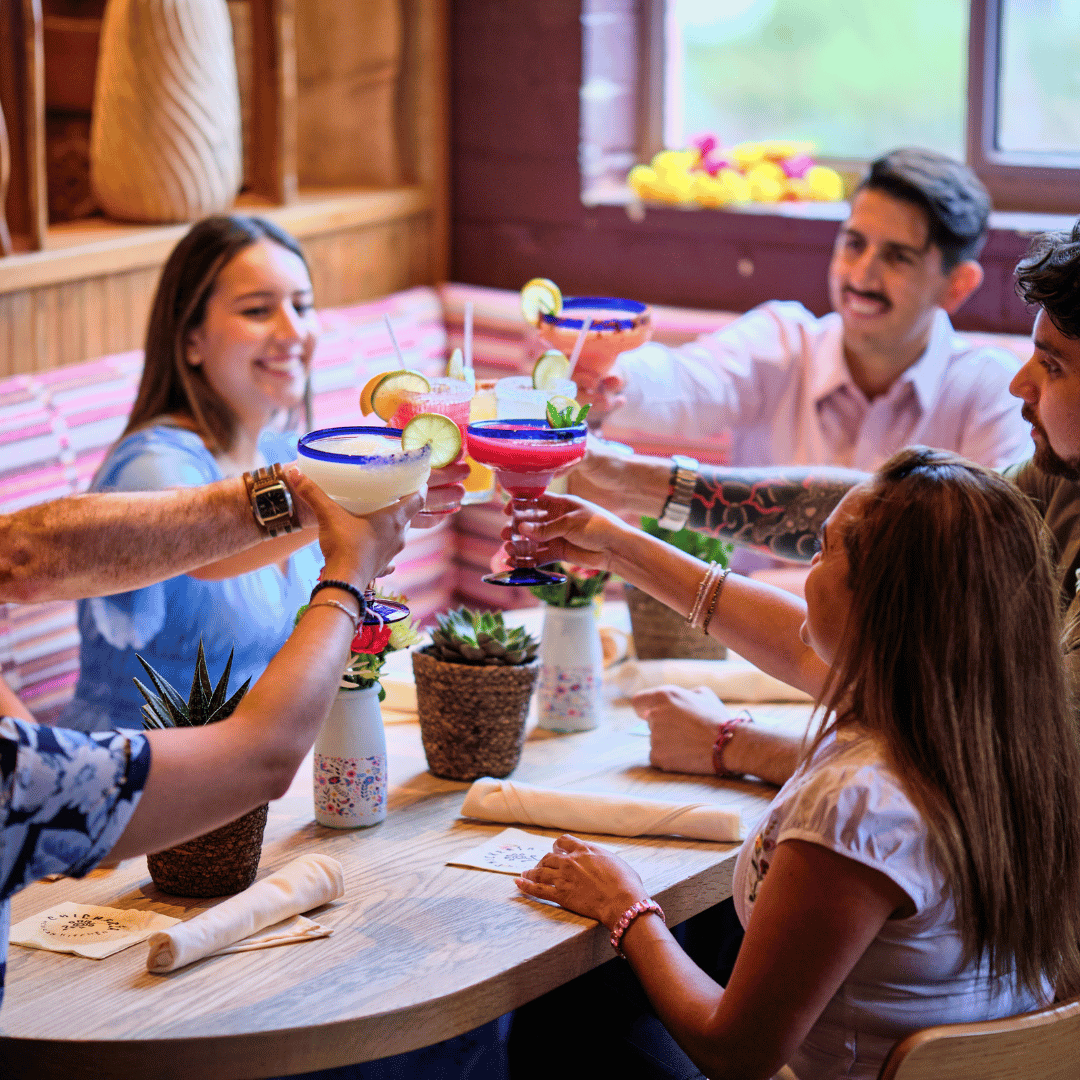People toasting with colorful cocktails at a celebration in a cozy, decorated restaurant with wooden walls and potted plants.
